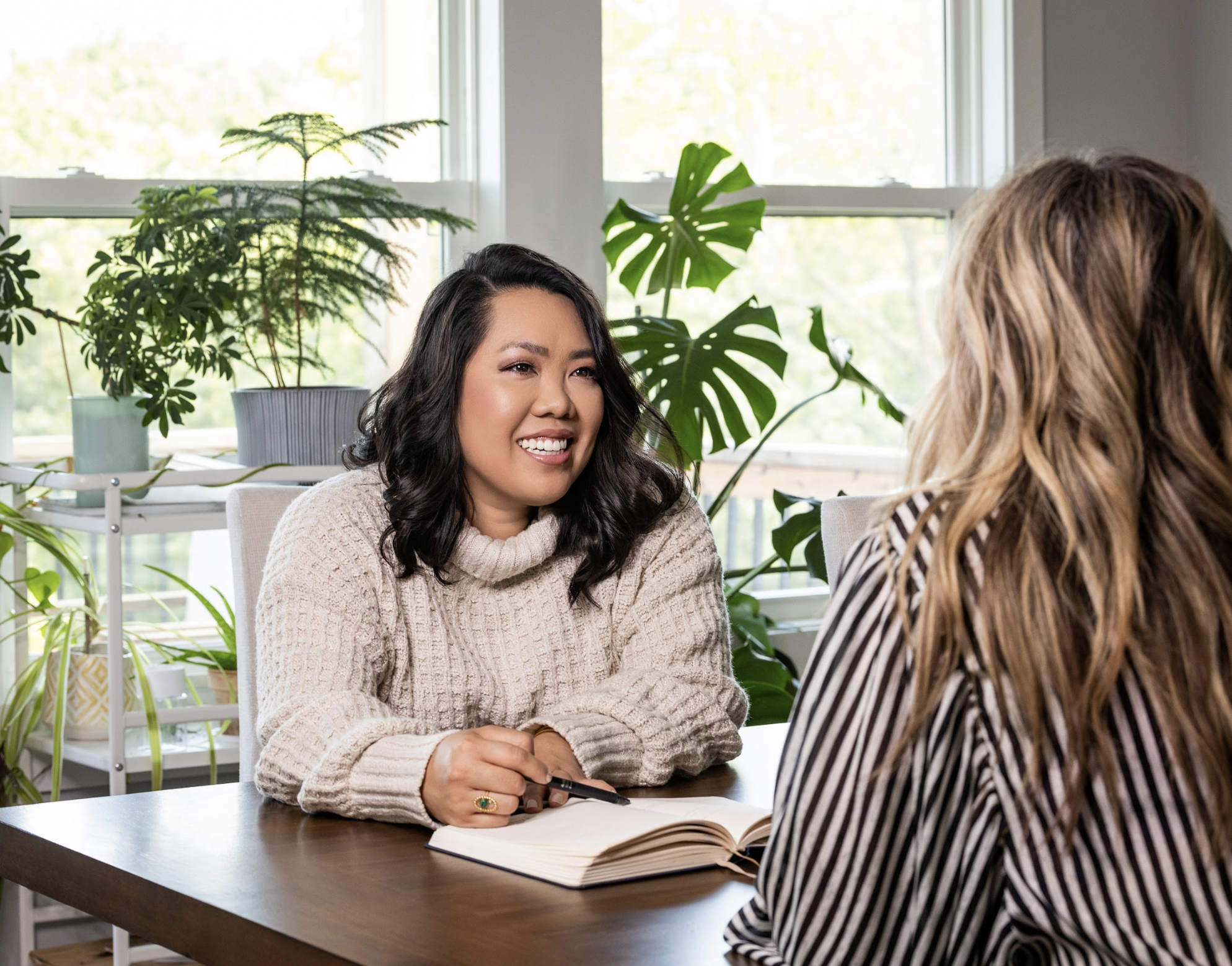 Two women are sitting at a wooden table in a well-lit room with large windows and various green plants in the background, engaged in a conversation. The woman on the left, with dark wavy hair, is smiling and holding a pen and an open notebook. The woman on the right has long, light brown hair with blonde highlights and is wearing a striped shirt.