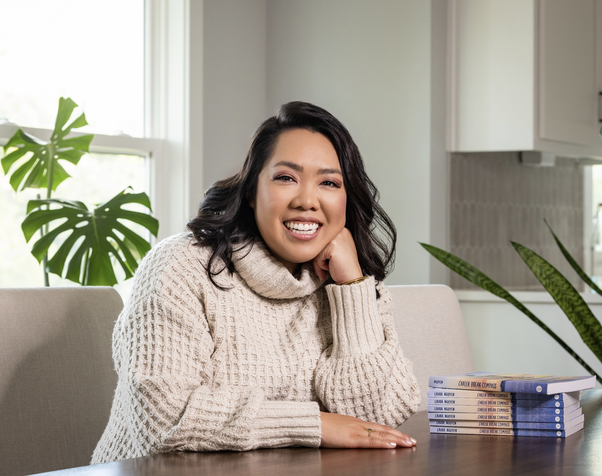 A woman sitting at a table in a well-lit room, smiling, with a stack of books titled "Career Break Compass" in front of her, and houseplants in the background.