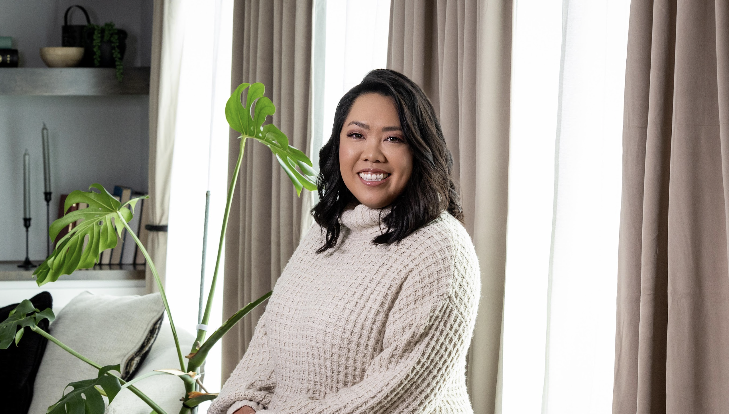 A woman with shoulder-length wavy dark hair, wearing a beige textured sweater, smiling and sitting near a window with beige curtains inside a living room with houseplants and candles.