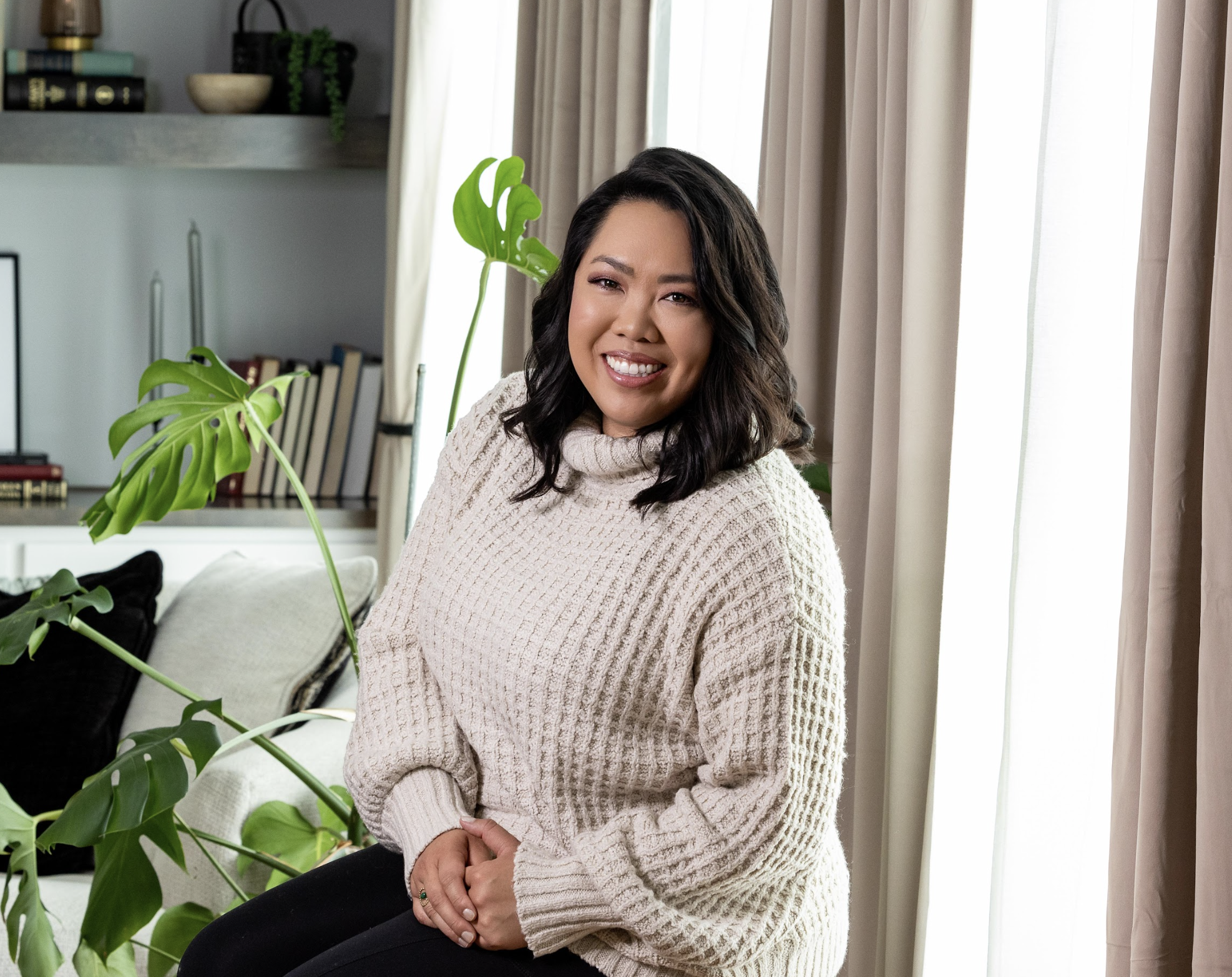 A woman with dark hair smiling while sitting on a sofa next to large indoor plants in a well-lit living room.
