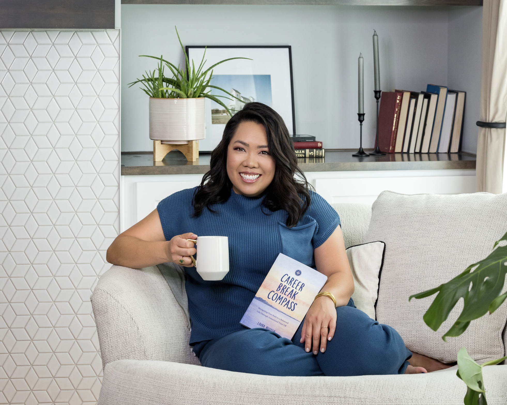 A smiling woman sitting on a beige couch in a living room, holding a white mug and a book titled "Career Break Compass." She has dark wavy hair and is wearing a blue top and blue pants.