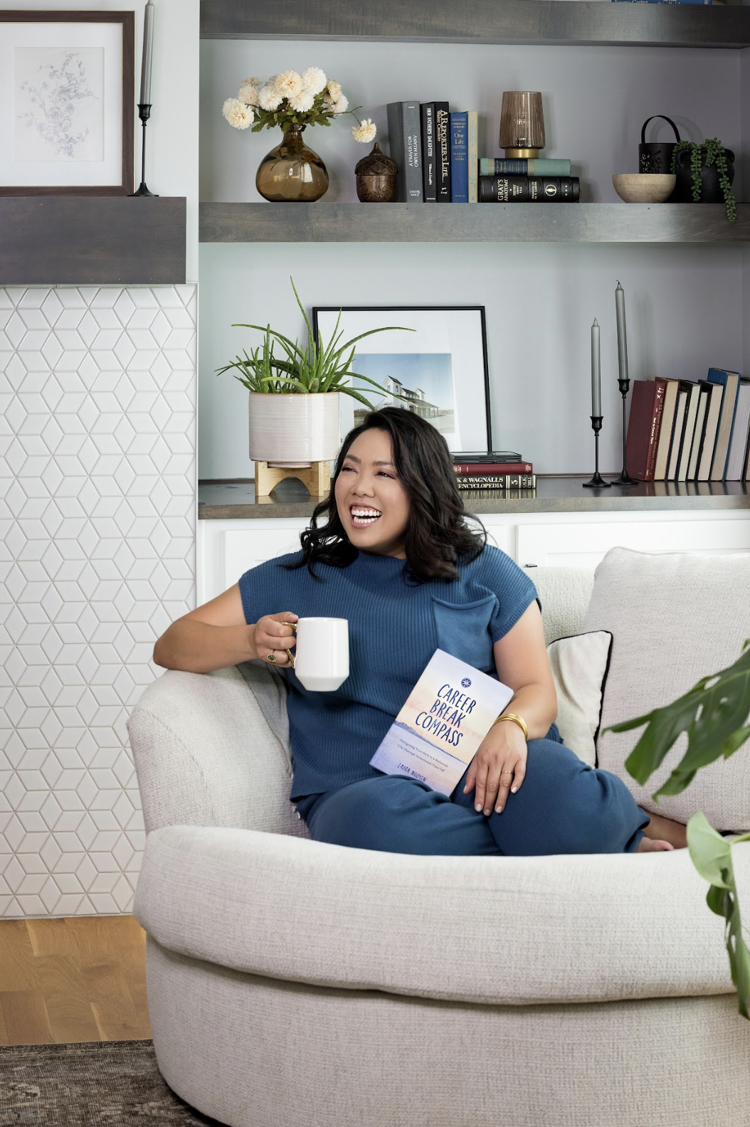 A woman sitting on a light-colored sofa in a living room, holding a white mug and a book titled "Career Break Compass", smiling and laughing. She is wearing a blue top and pants, with indoor plants and bookshelves in the background.