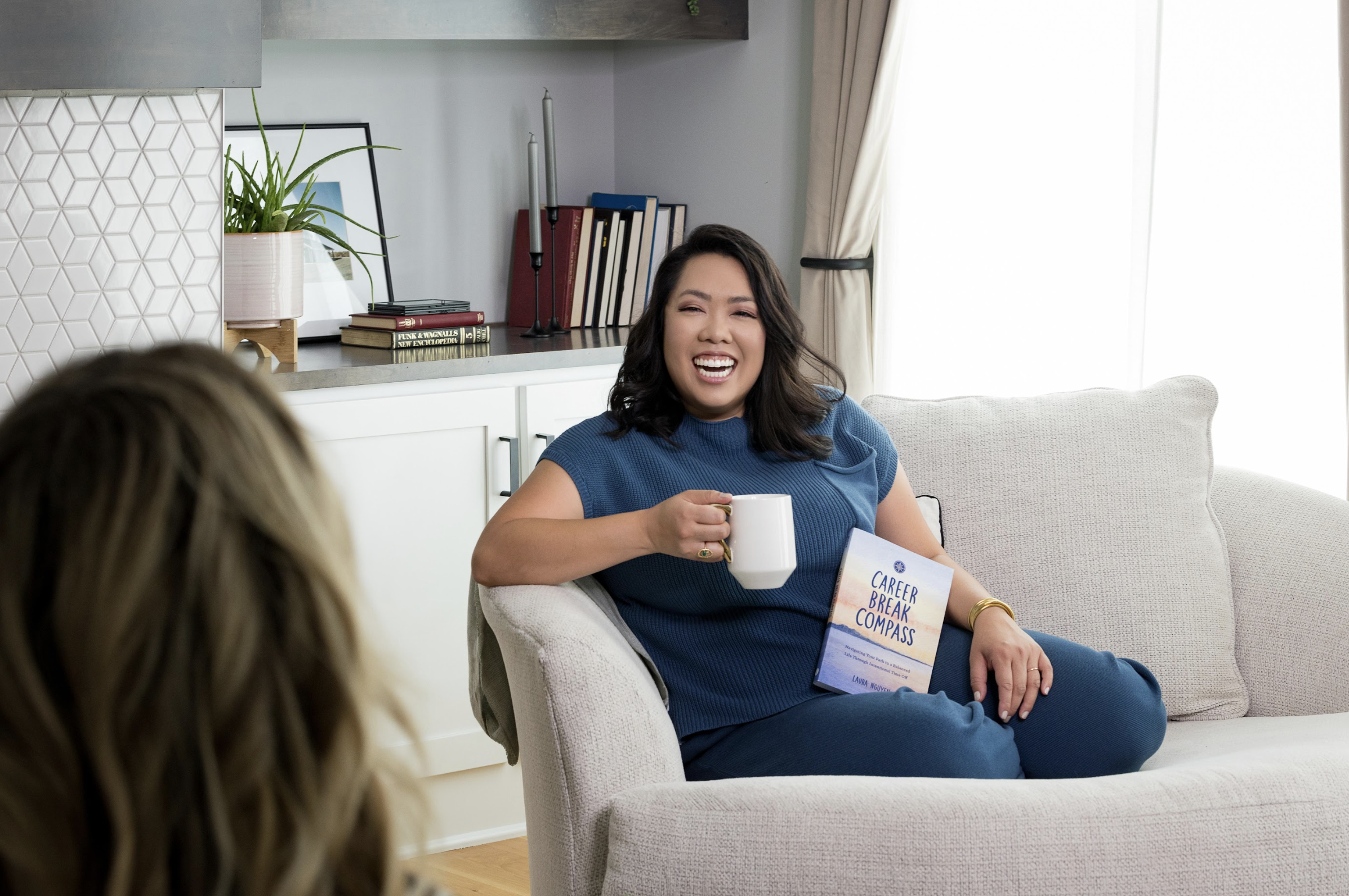 Woman sitting on a sofa holding a book titled 'Career Break Compass' and a mug, smiling and talking to another person in a cozy living room.