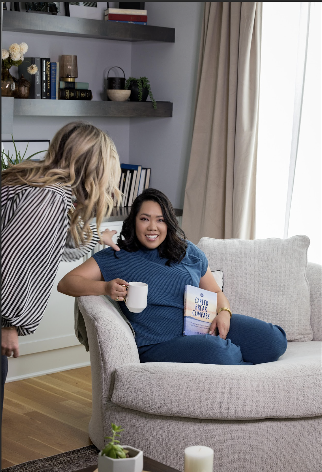 A woman sitting on a beige sofa in a living room, holding a white mug and a book titled 'Career Break Compass', smiling at another woman leaning towards her. The room has a bookshelf and decorative items on the wall behind them, and a window with beige curtains letting in natural light.