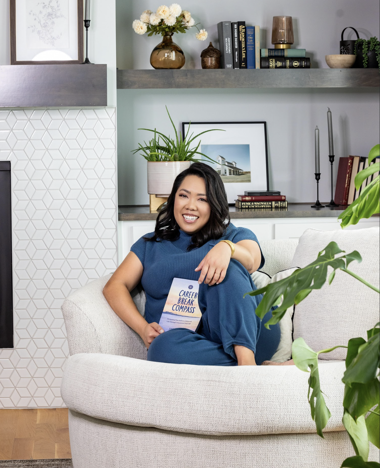 A woman sitting on a white couch in a modern living room, holding a book titled 'Career Break Compass', smiling at the camera. The background features a shelf with books, a framed photo, and decorative items, along with plants and a lamp.