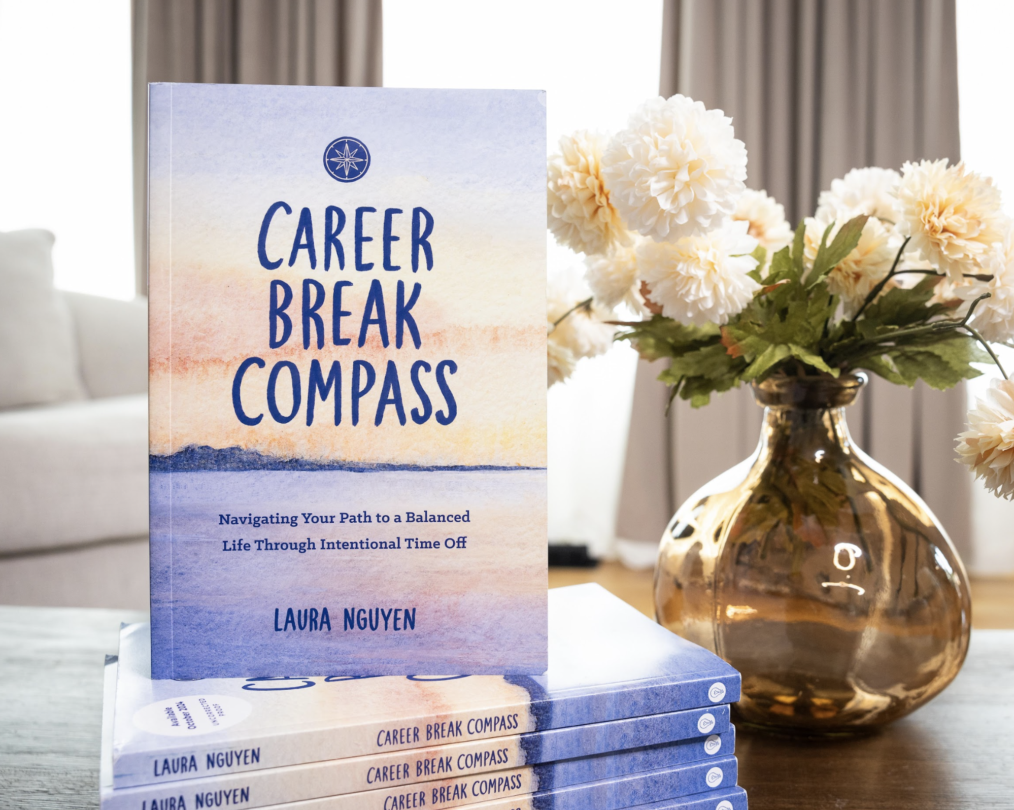 A stack of books titled 'Career Break Compass' by Laura Nguyen on a table, next to a glass vase with white flowers, in a well-lit room with curtains in the background.