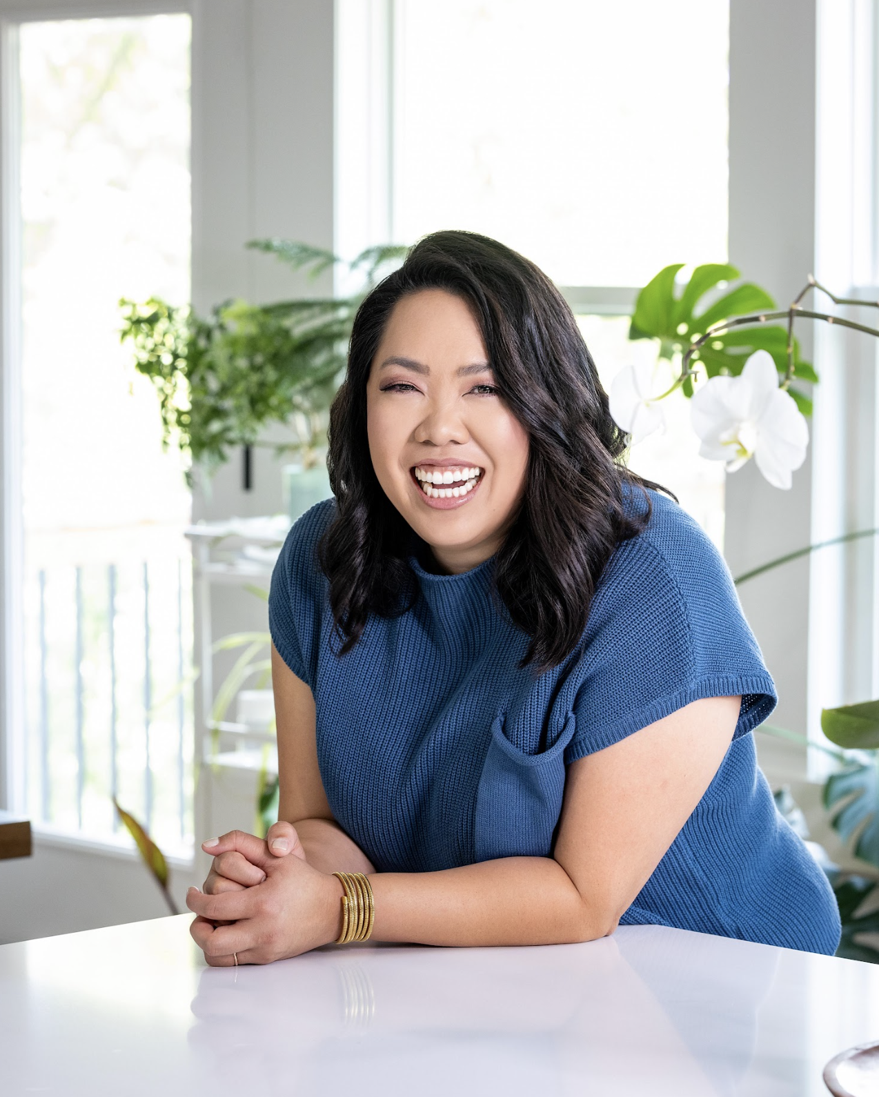 A woman with black wavy hair smiling and leaning on a white table, wearing a blue top, with green plants and orchids in the background.