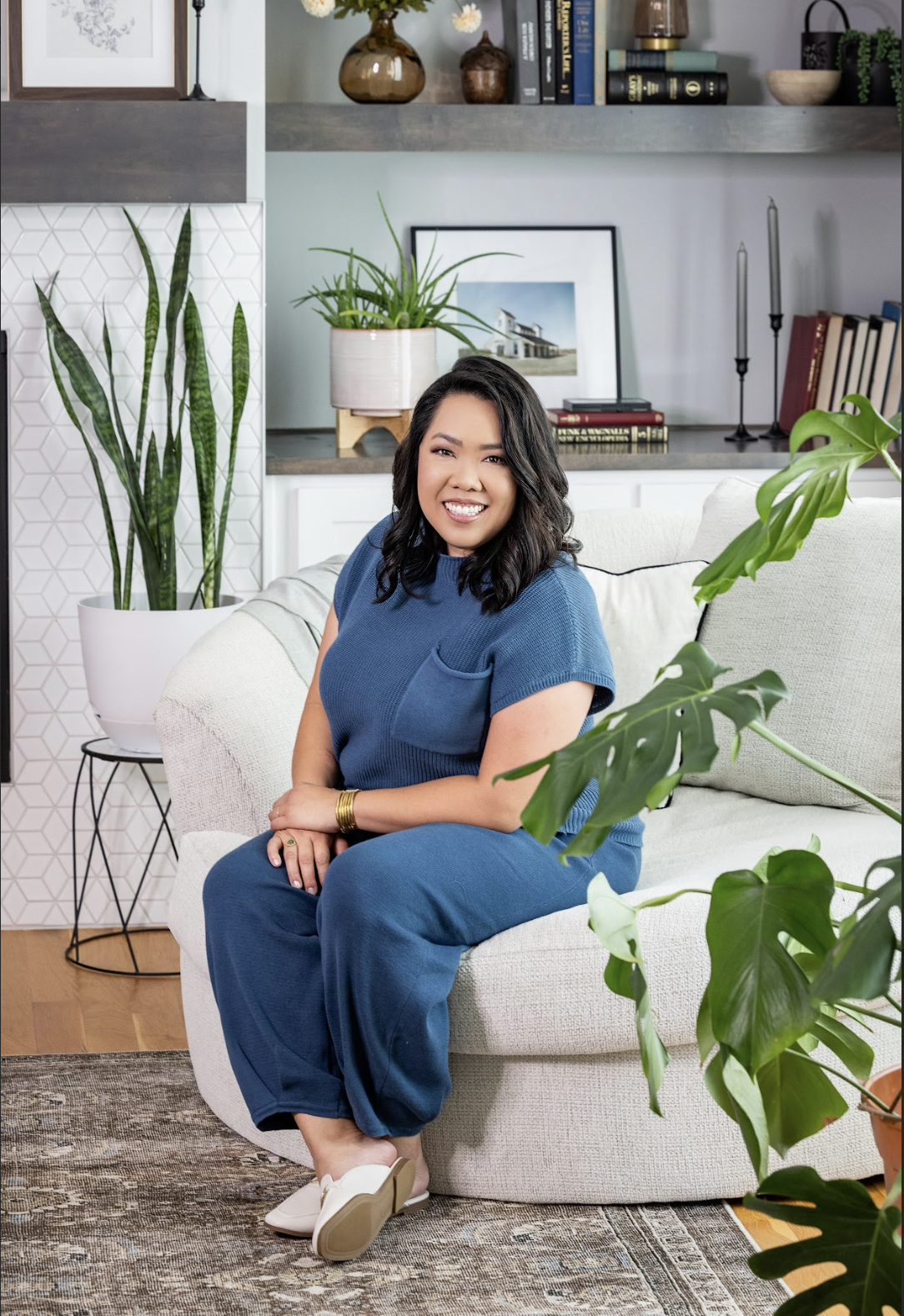 A woman with dark, wavy hair sitting on a light-colored couch in a modern living room, smiling at the camera. She is wearing a blue outfit, beige shoes, and gold jewelry. The background includes green potted plants, a white and gray wall with shelving, framed artwork, books, and decorative objects.