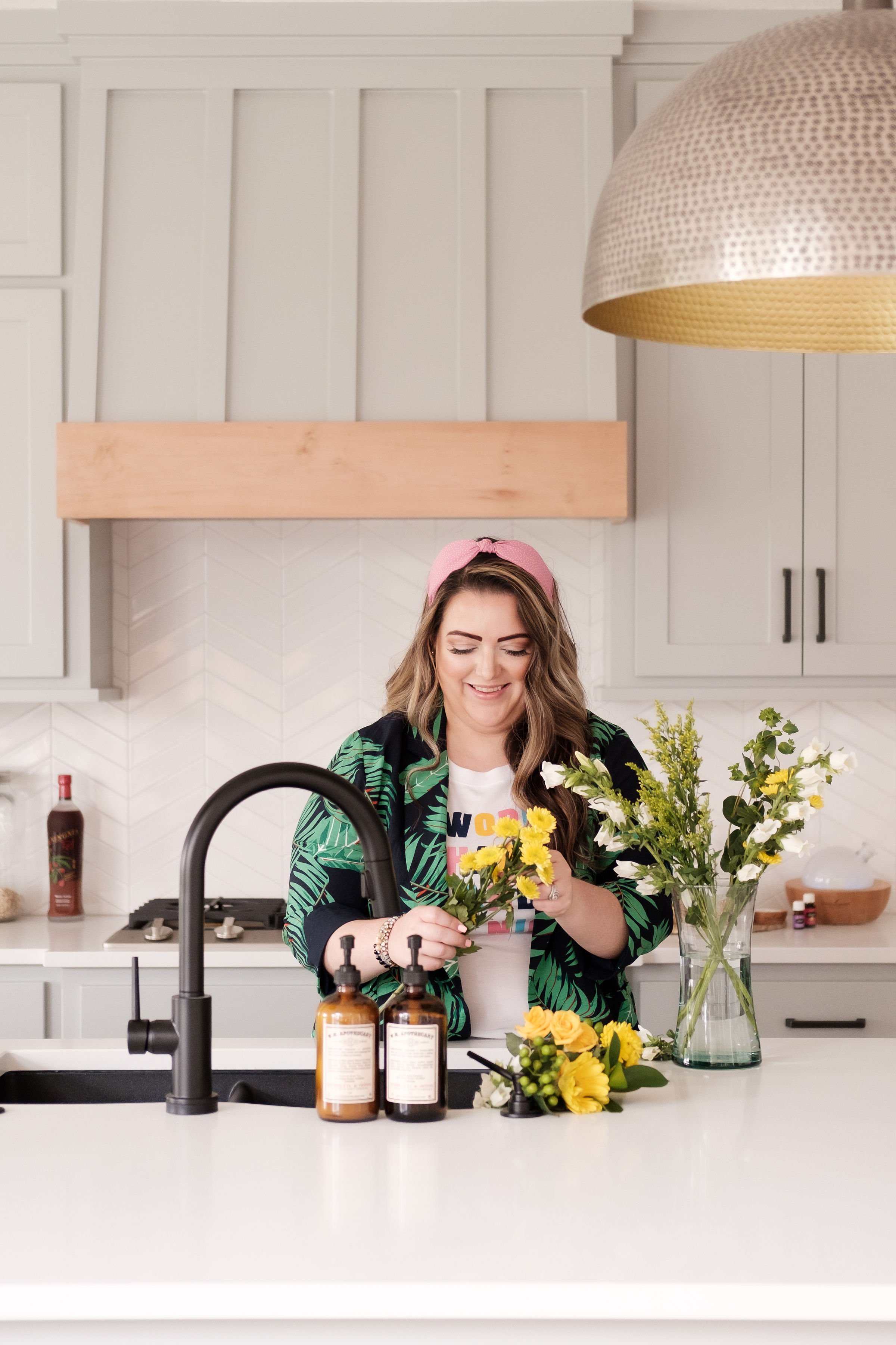 A woman with long wavy hair, wearing a pink headband, a graphic t-shirt, and a tropical print jacket, is smiling while arranging yellow and white flowers in a kitchen with white cabinets and a countertop.
