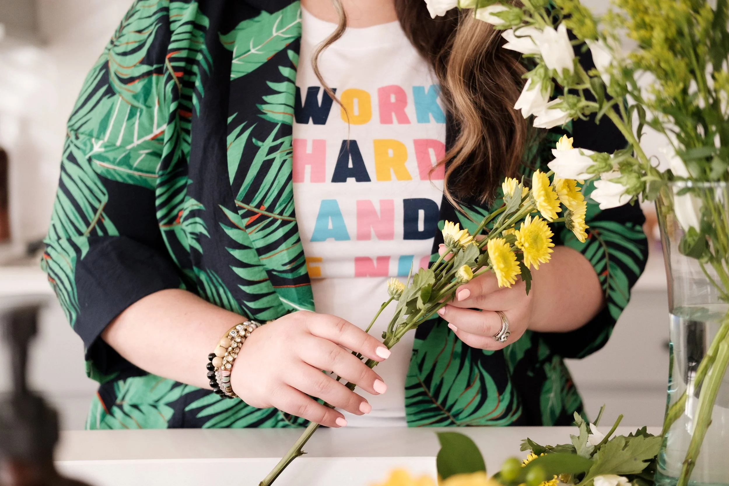 Person arranging yellow and white flowers in a vase, wearing a white T-shirt with multicolored text and a green leafy patterned jacket.