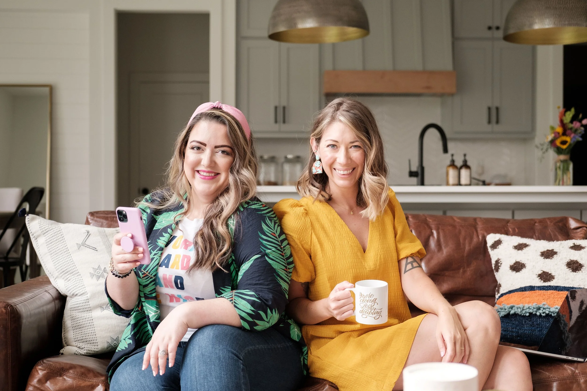 Two women sitting on a brown leather couch in a modern kitchen, smiling and enjoying coffee and a phone.