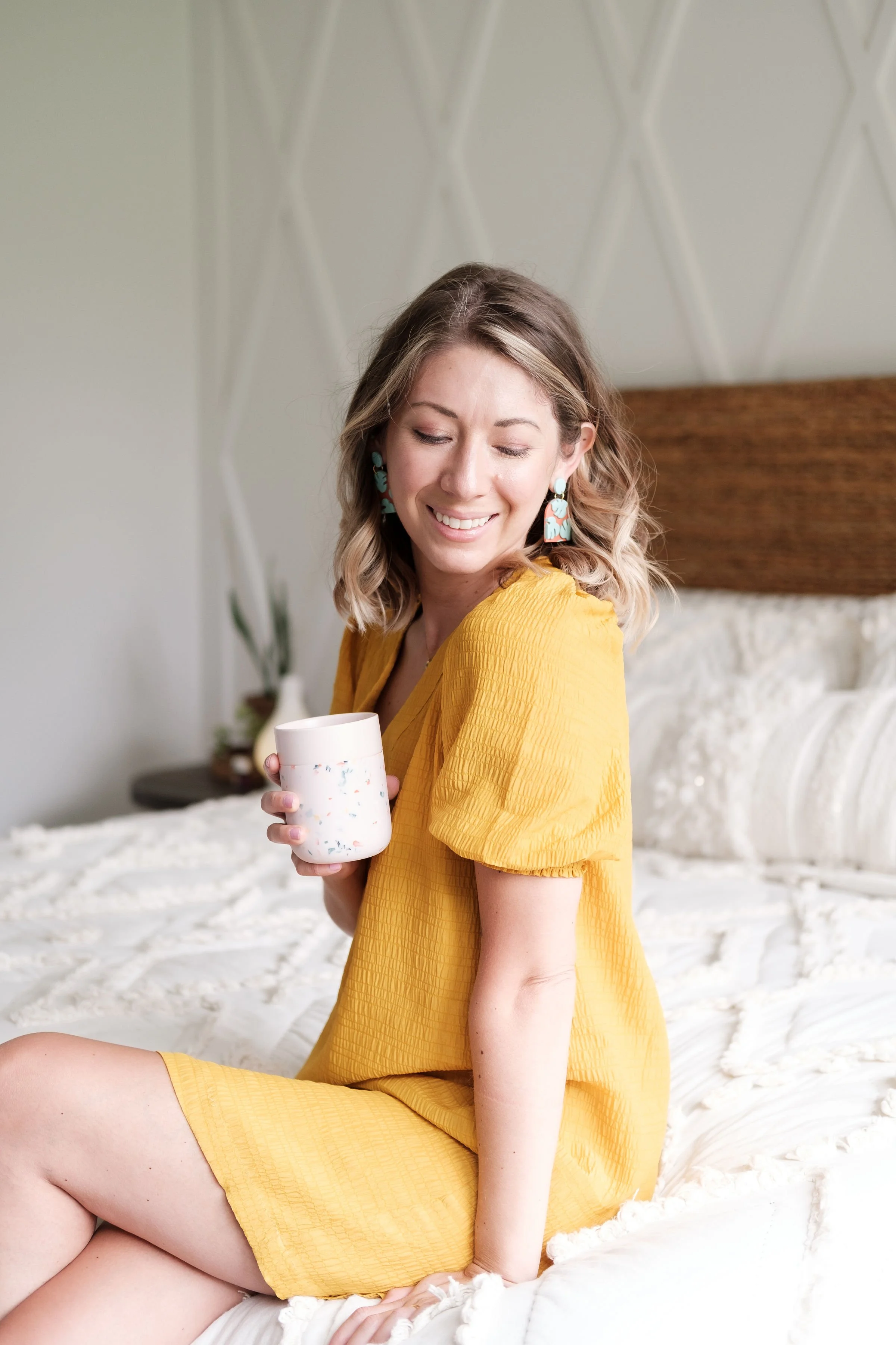 A woman wearing a yellow dress sitting on a bed, holding a white cup with colorful specks, smiling with eyes closed.