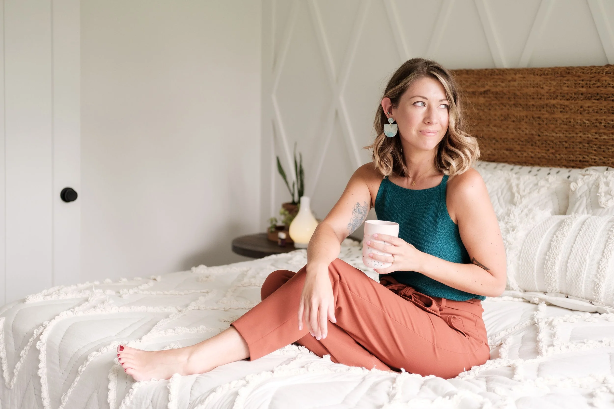 A woman with wavy blonde hair sitting on a bed, holding a white mug, wearing a teal sleeveless top and rust-colored pants, looking to the side with a slight smile in a bedroom with a wooden headboard and white textured bedding.