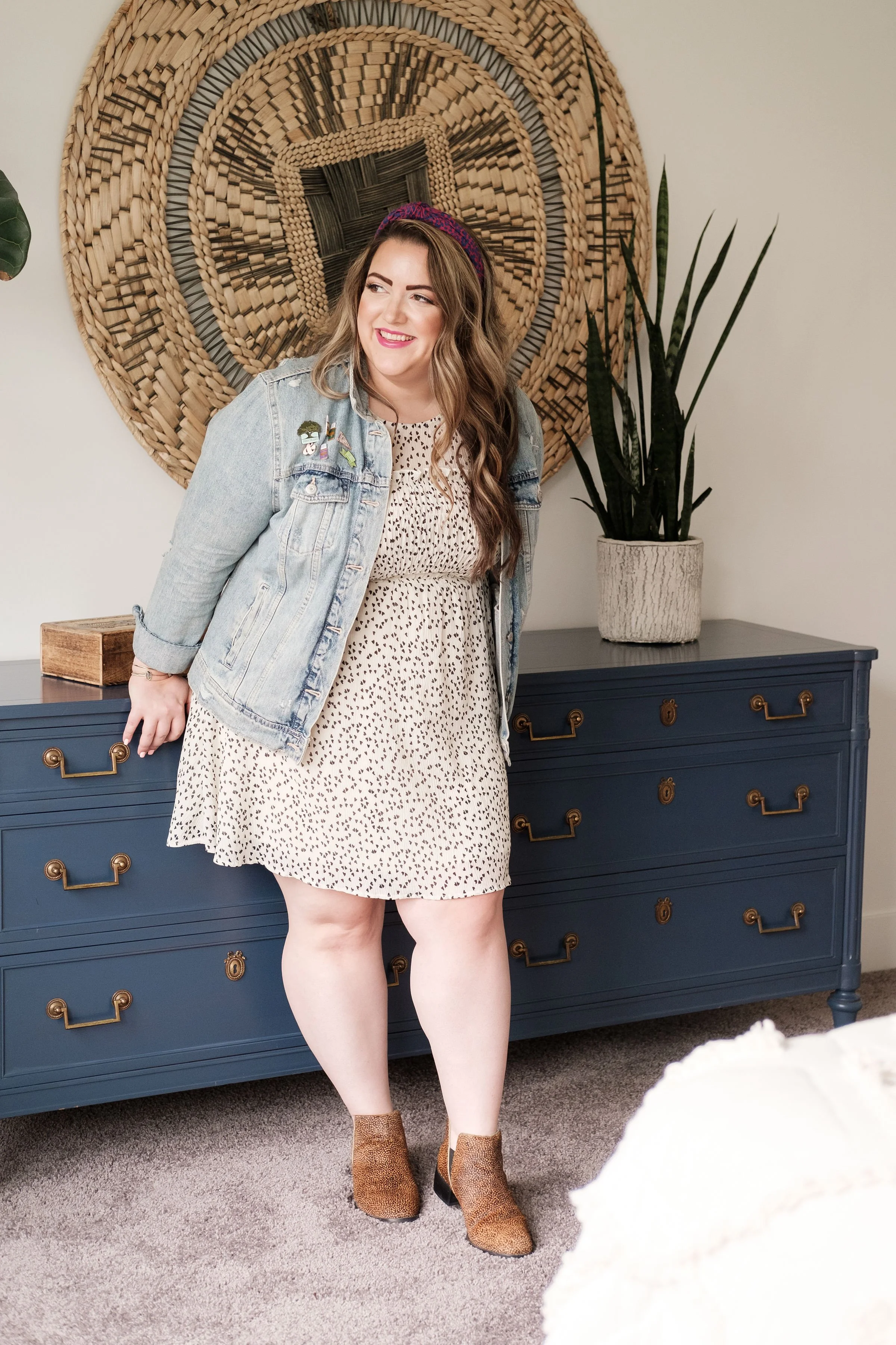 A woman with long wavy hair, wearing a denim jacket with patches, a leopard print dress, and brown ankle boots, standing in a room with a blue dresser, a large woven wall hanging, and a tall potted plant.