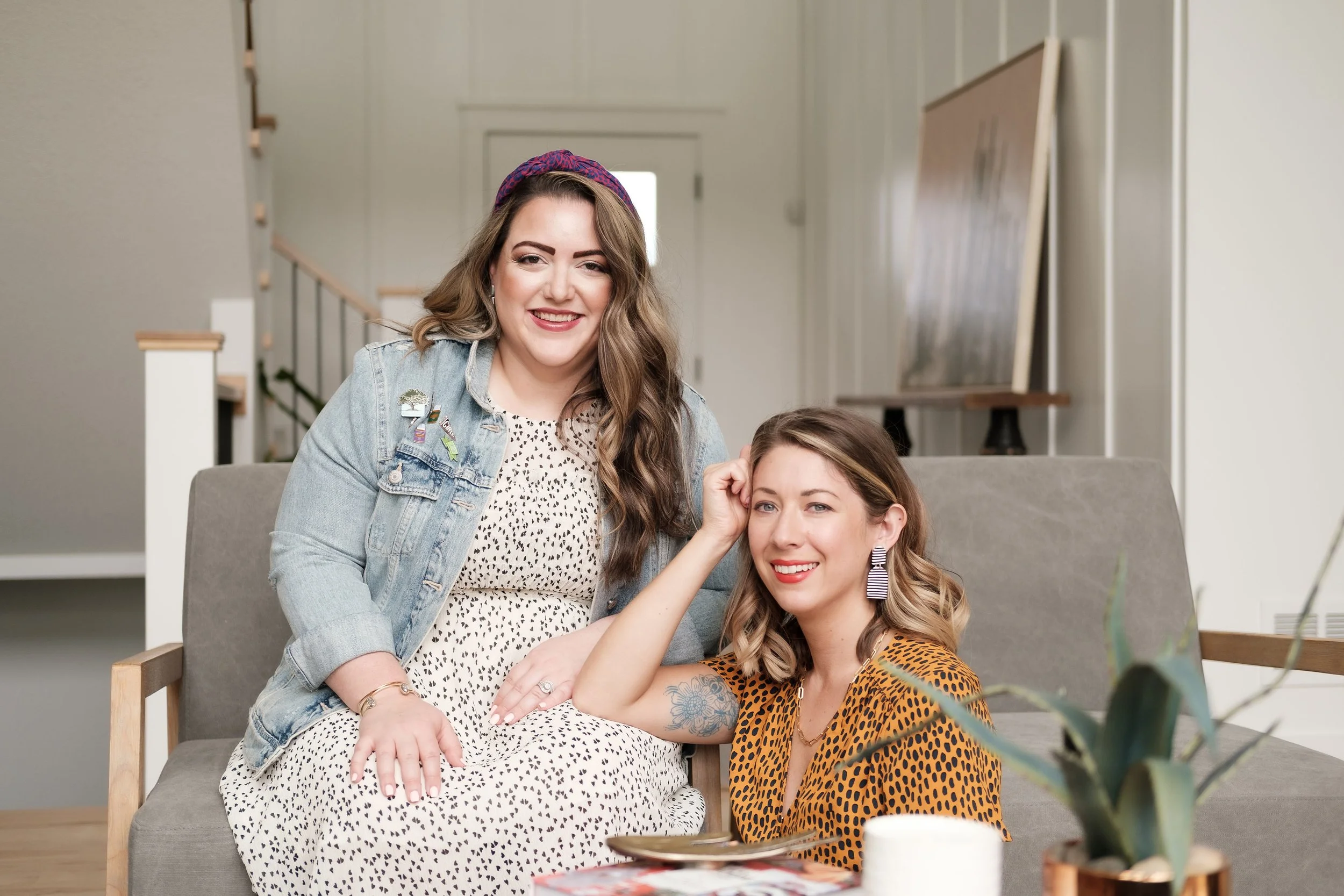 Two women sitting on a sofa in a bright living room, smiling at the camera. One is wearing a denim jacket and a patterned dress, the other is wearing an animal print top and earrings, with a tattoo on her arm. There is a plant and a roll of paper towels on a table in front of them.