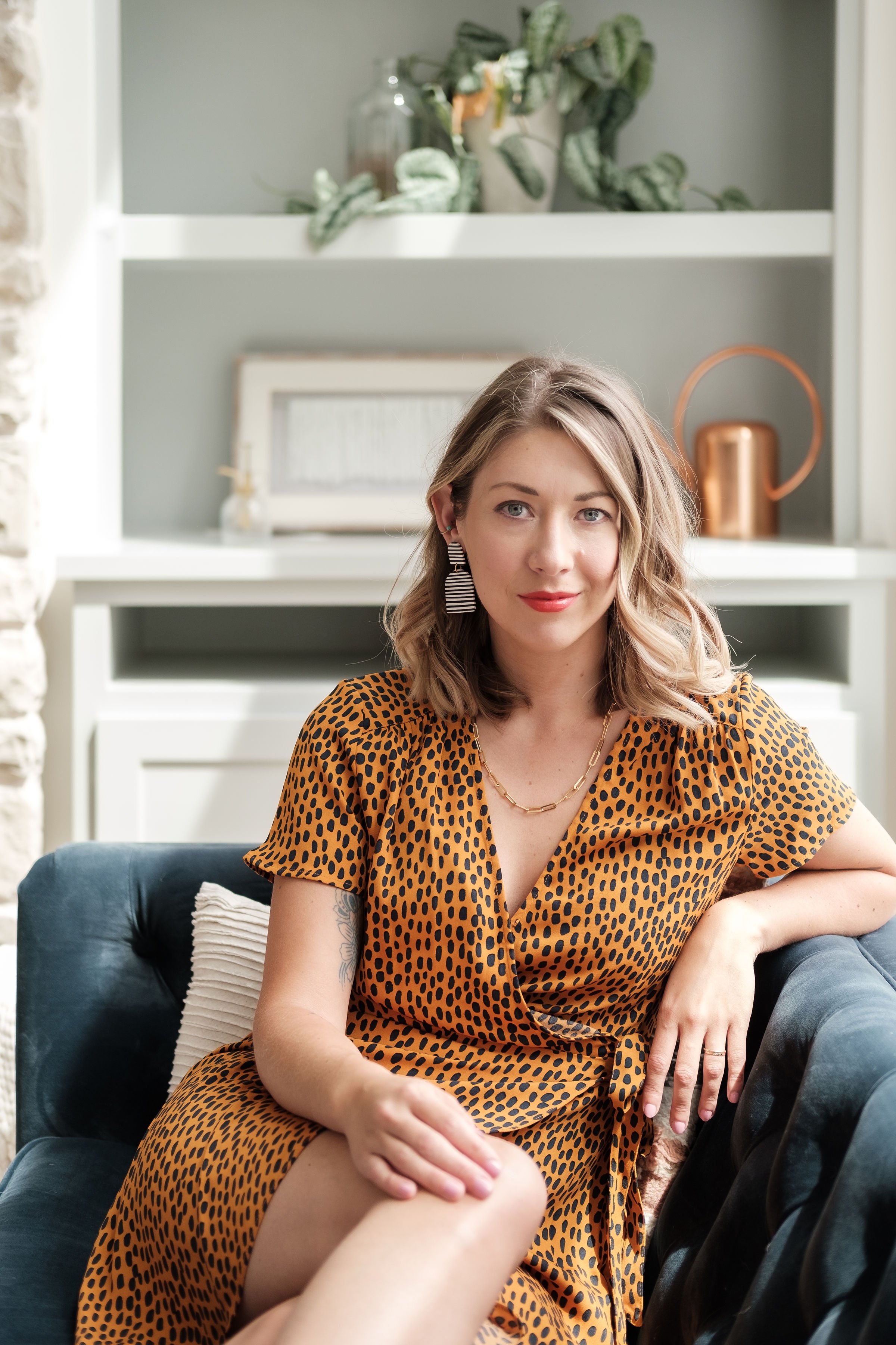 A woman with blonde hair wearing a yellow and black polka dot dress, sitting on a dark blue couch, inside a cozy room with decorative shelves and plants in the background.
