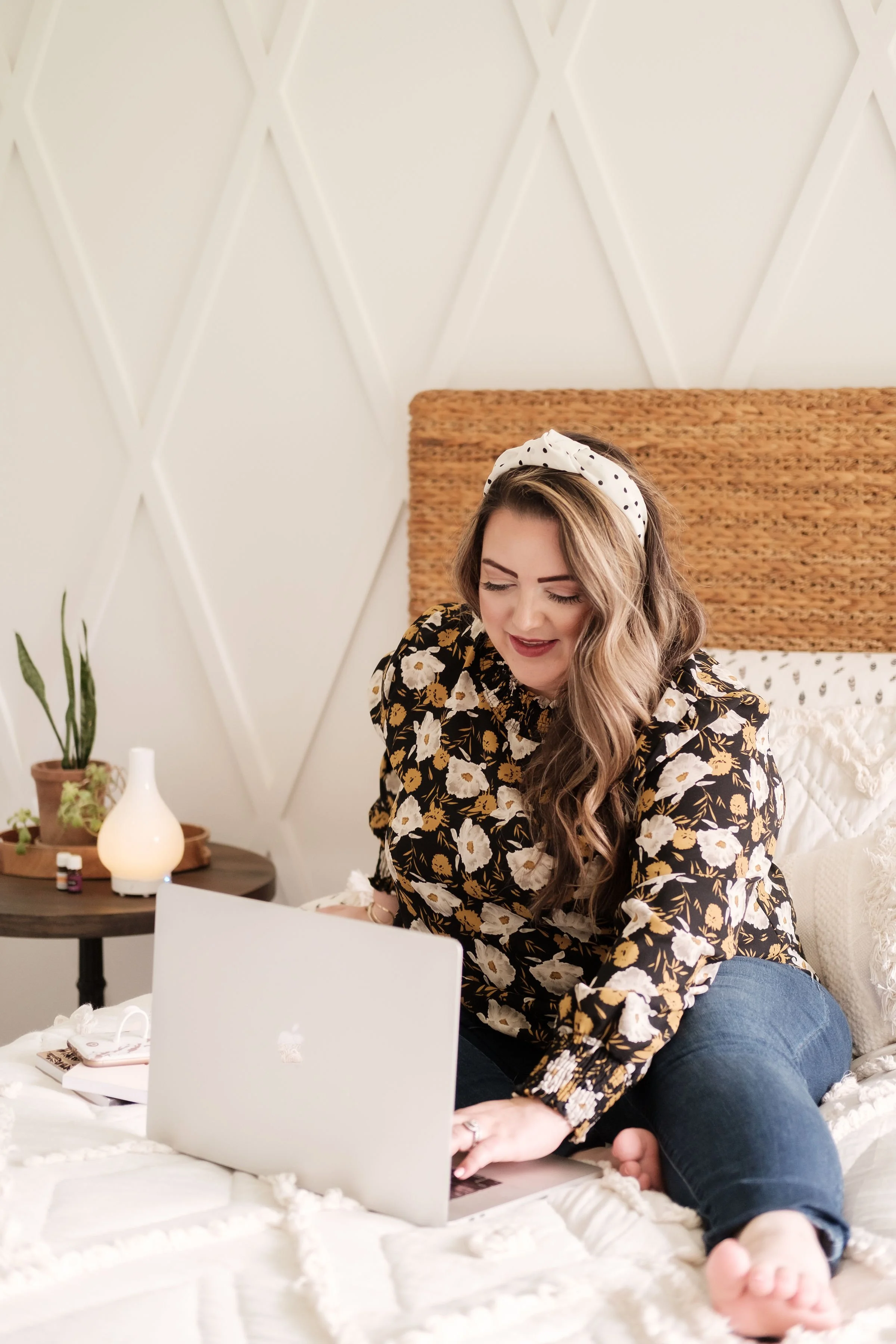 A woman with long wavy hair, wearing a floral blouse and a white polka dot headband, sitting on a bed with a white quilt, using a silver MacBook laptop.