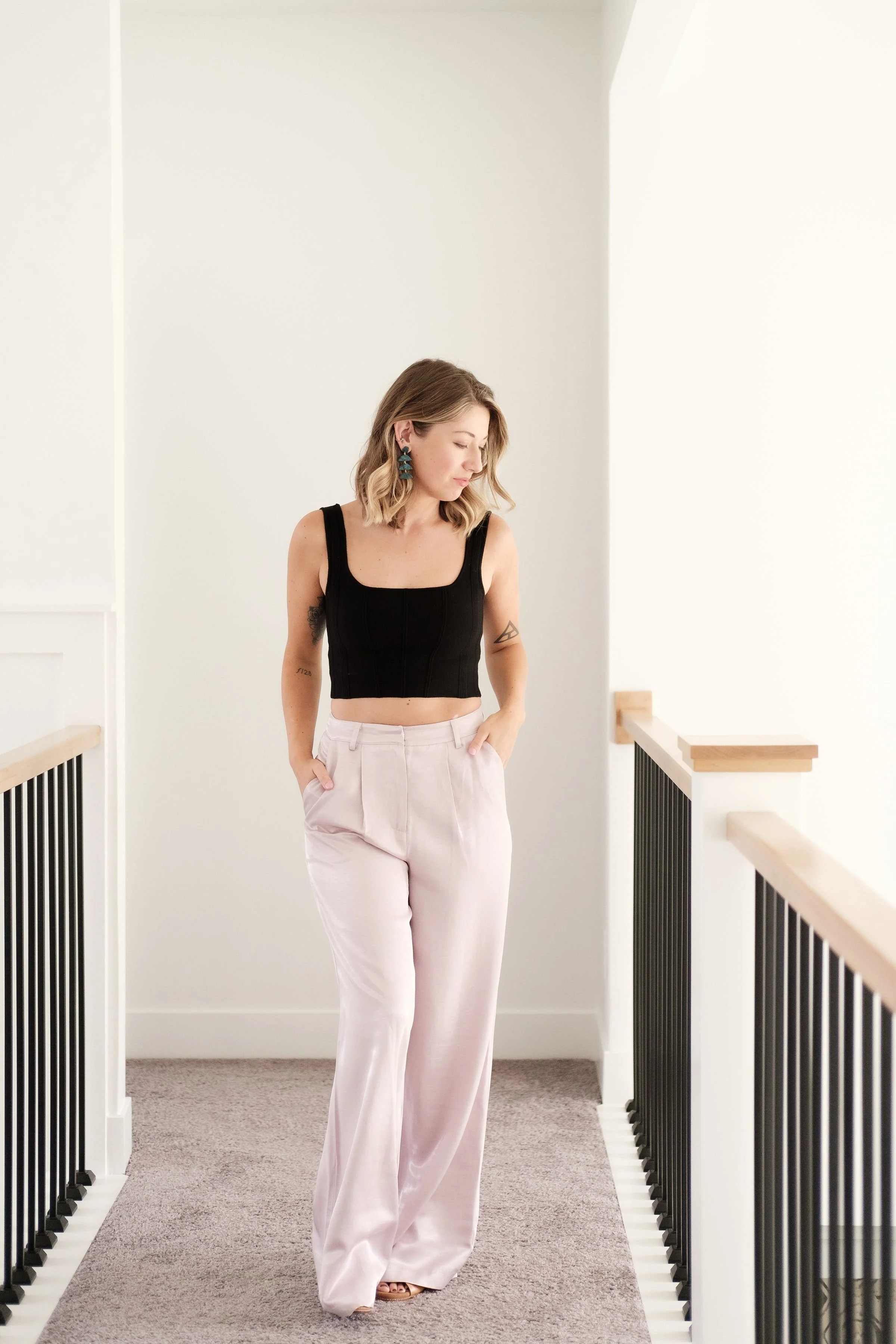 A woman walking down a narrow, carpeted hallway with white walls and black railing, wearing a black crop top, light pink wide-leg pants, and turquoise earrings.