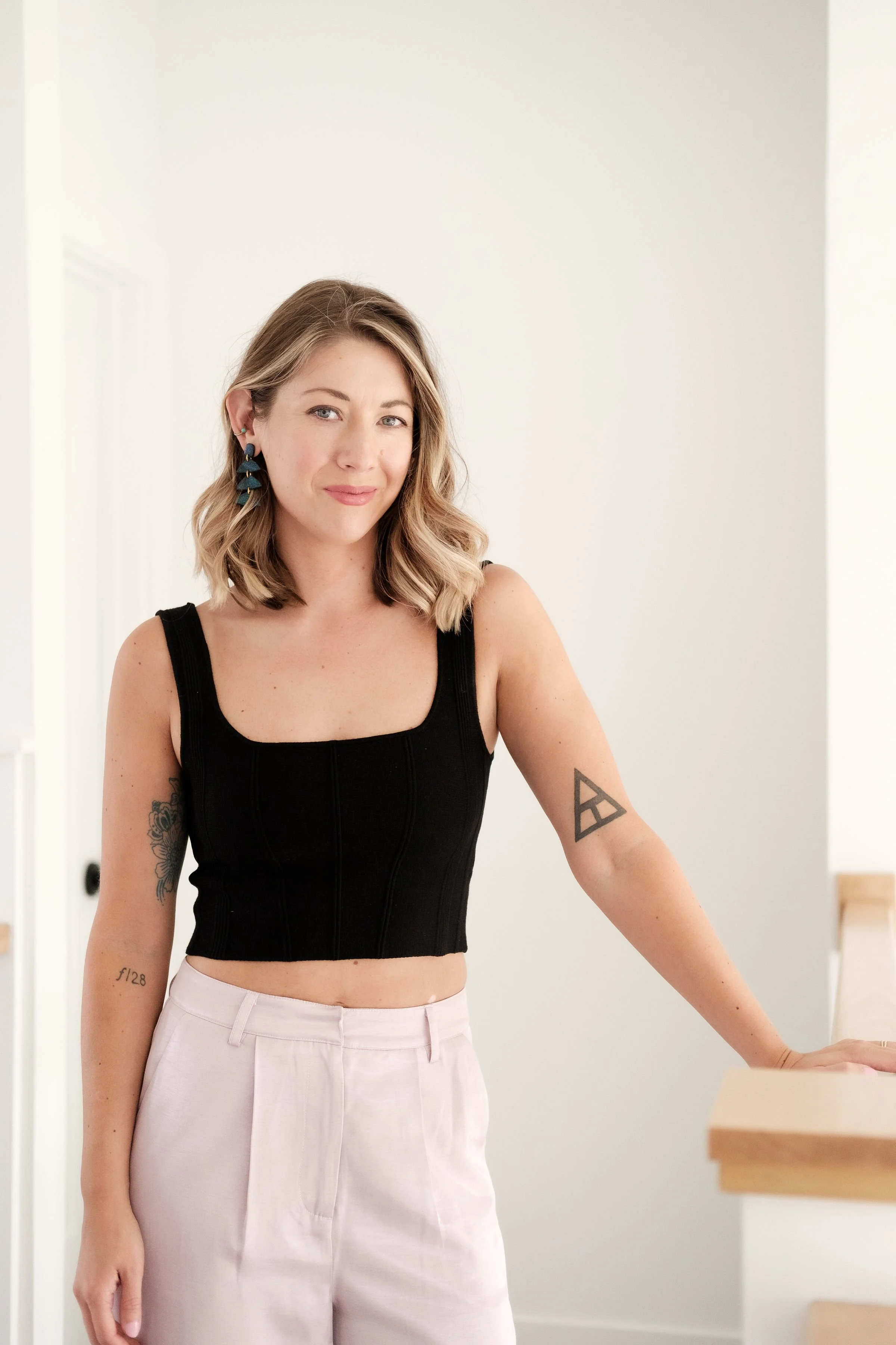 A woman with shoulder-length blonde hair and light skin, wearing a black sleeveless crop top and light-colored pants, standing inside a bright room with white walls and a wooden railing, smiling slightly.