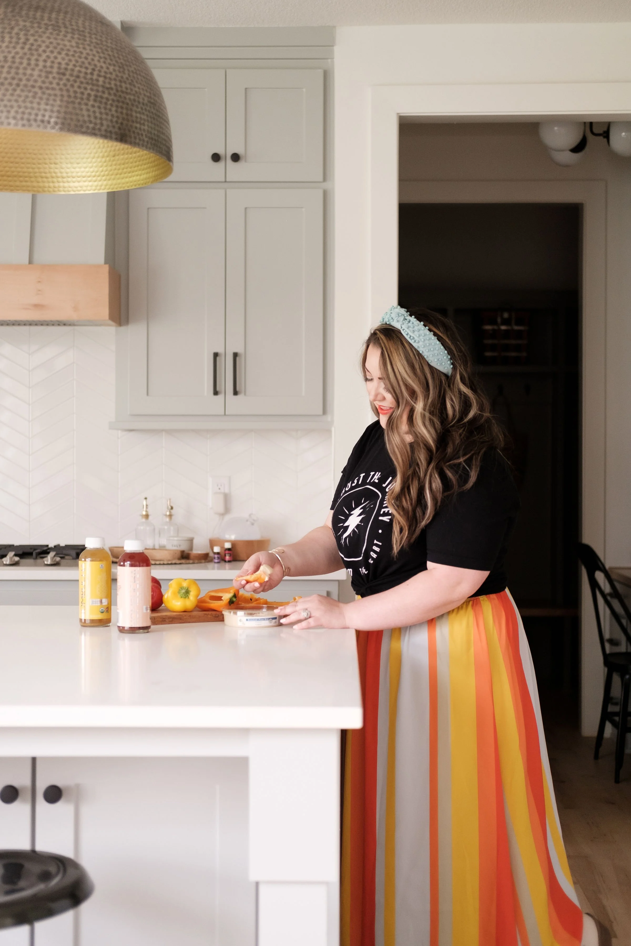 A woman with long wavy hair wearing a black t-shirt and a colorful striped skirt is preparing food in a bright kitchen. She is standing at a white countertop with condiments and vegetables, smiling as she spreads a dip or spread on a piece of bread or cracker.