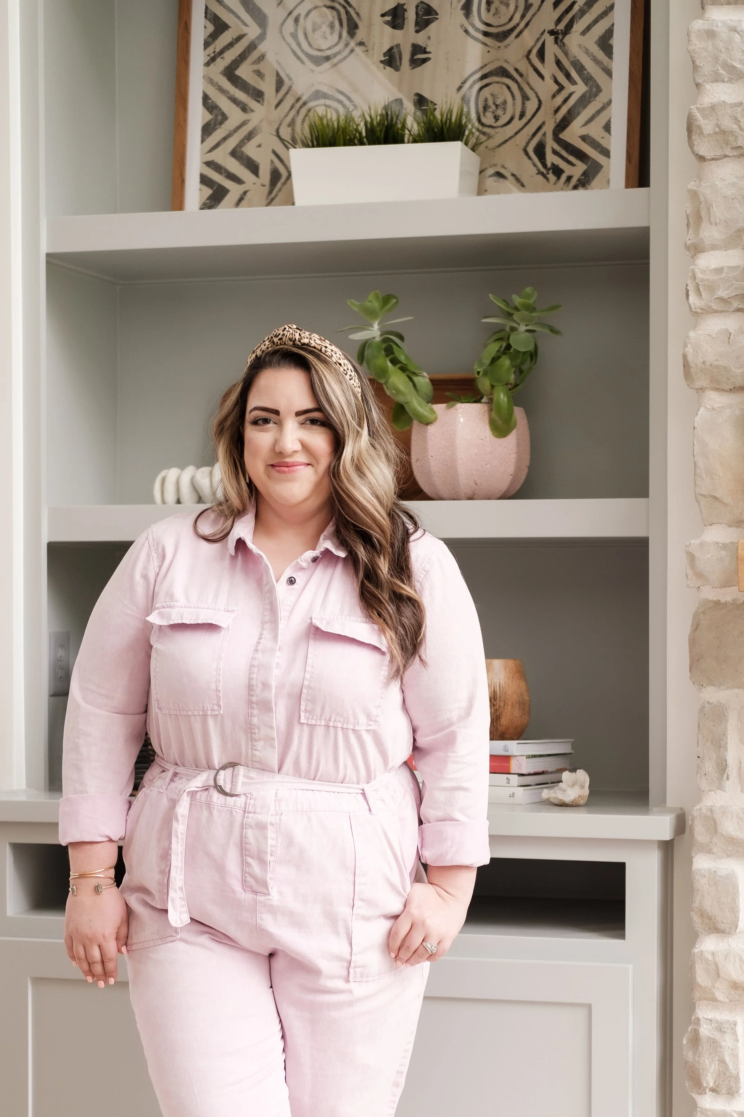 A woman with long, wavy hair wearing a light pink jumpsuit with a belt, standing in front of a white shelf with plants and books.