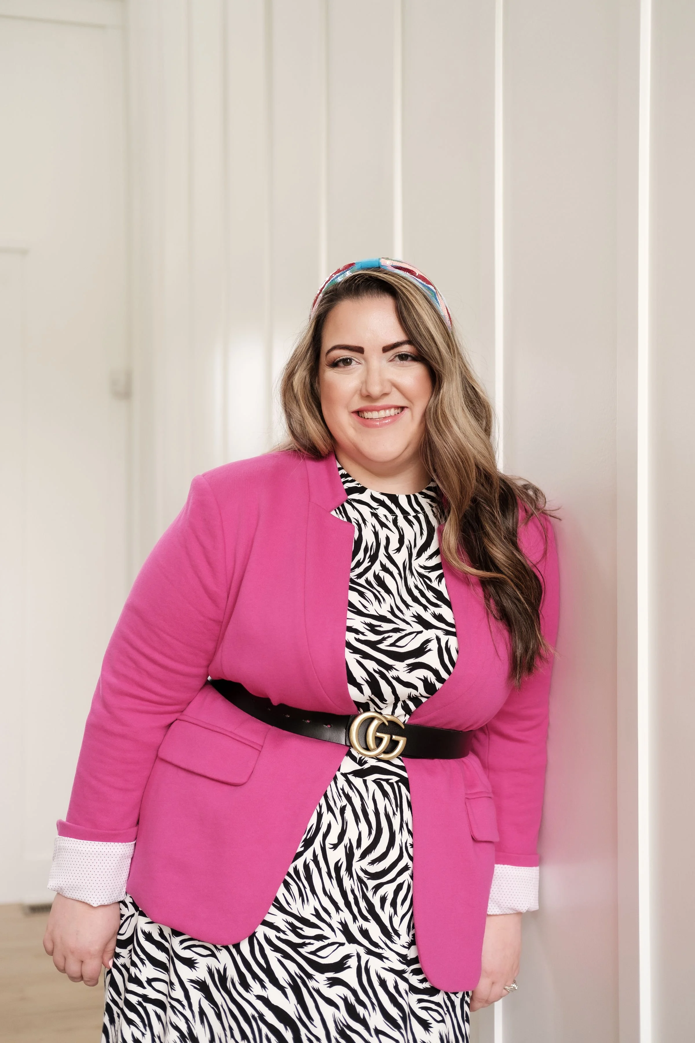 A woman with long wavy brown hair, wearing a pink blazer over a zebra print dress, black belt with gold GG buckle, and a colorful headband, standing against a white wall with a smile.