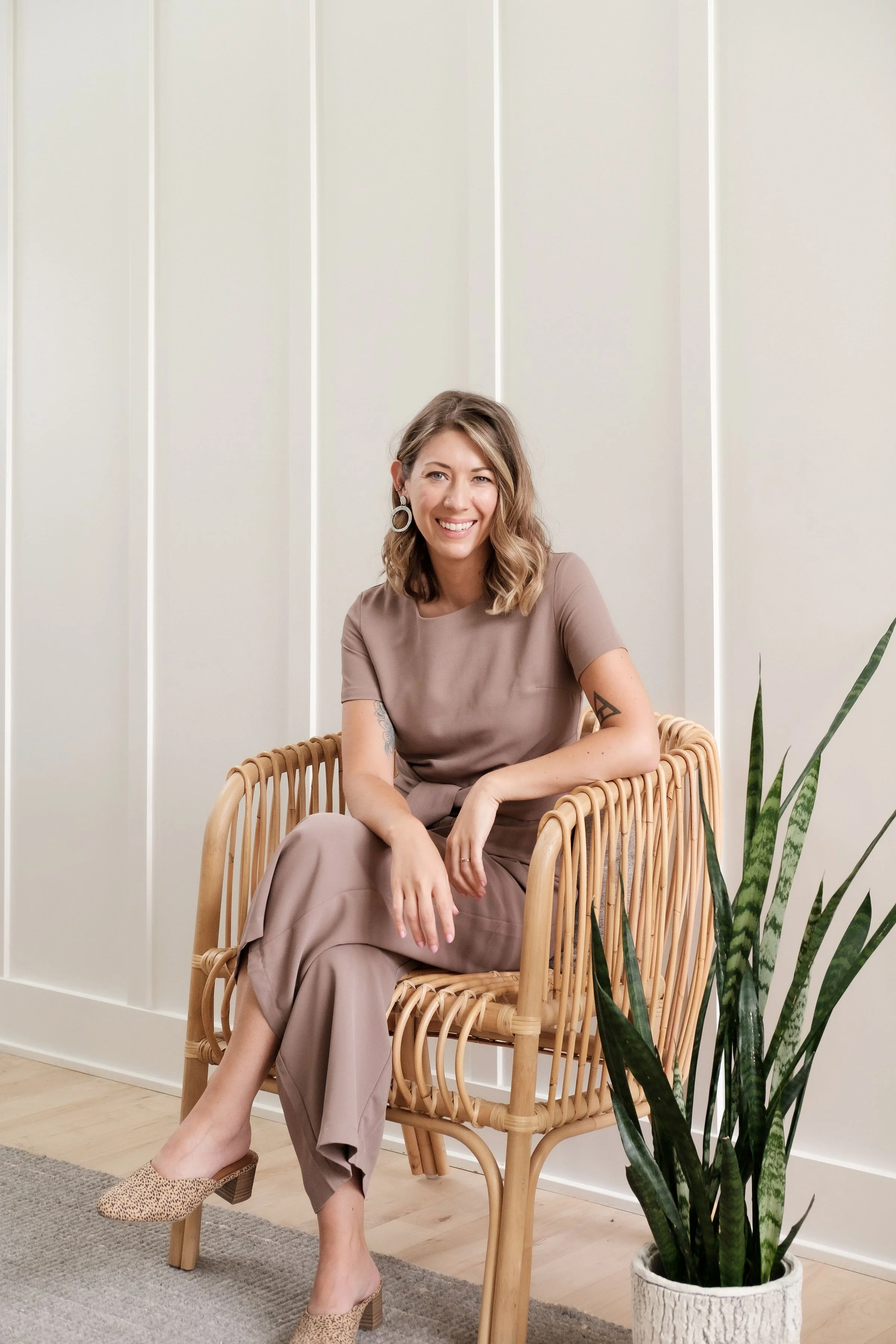 A smiling woman with shoulder-length wavy hair, wearing a taupe outfit, sitting on a rattan chair next to a potted snake plant on the floor in a minimalist room.