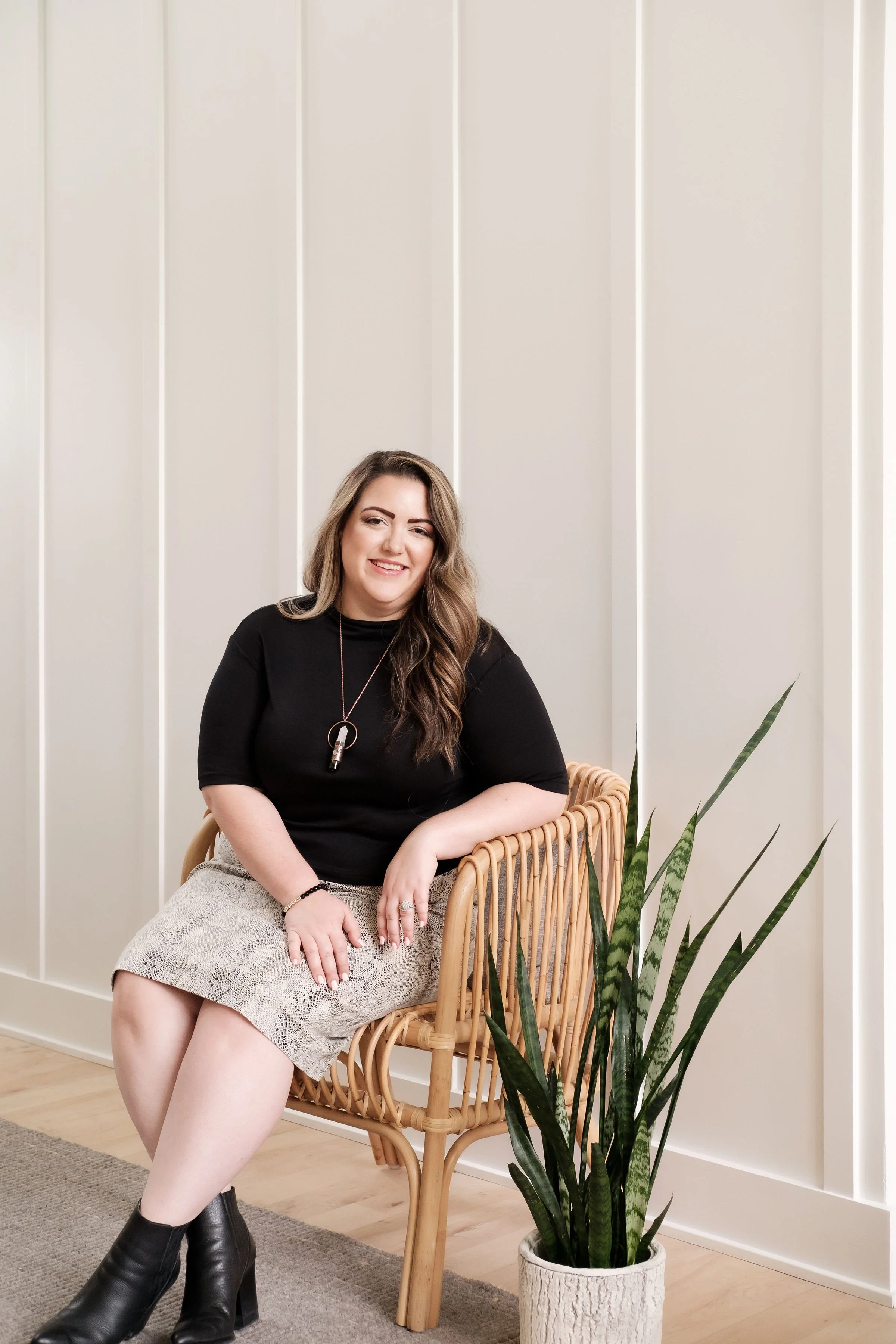 A woman with long brown hair wearing a black top, snake print skirt, and black ankle boots, sitting on a rattan chair next to a large plant in a white textured pot, in a room with white paneled walls and light wood flooring.