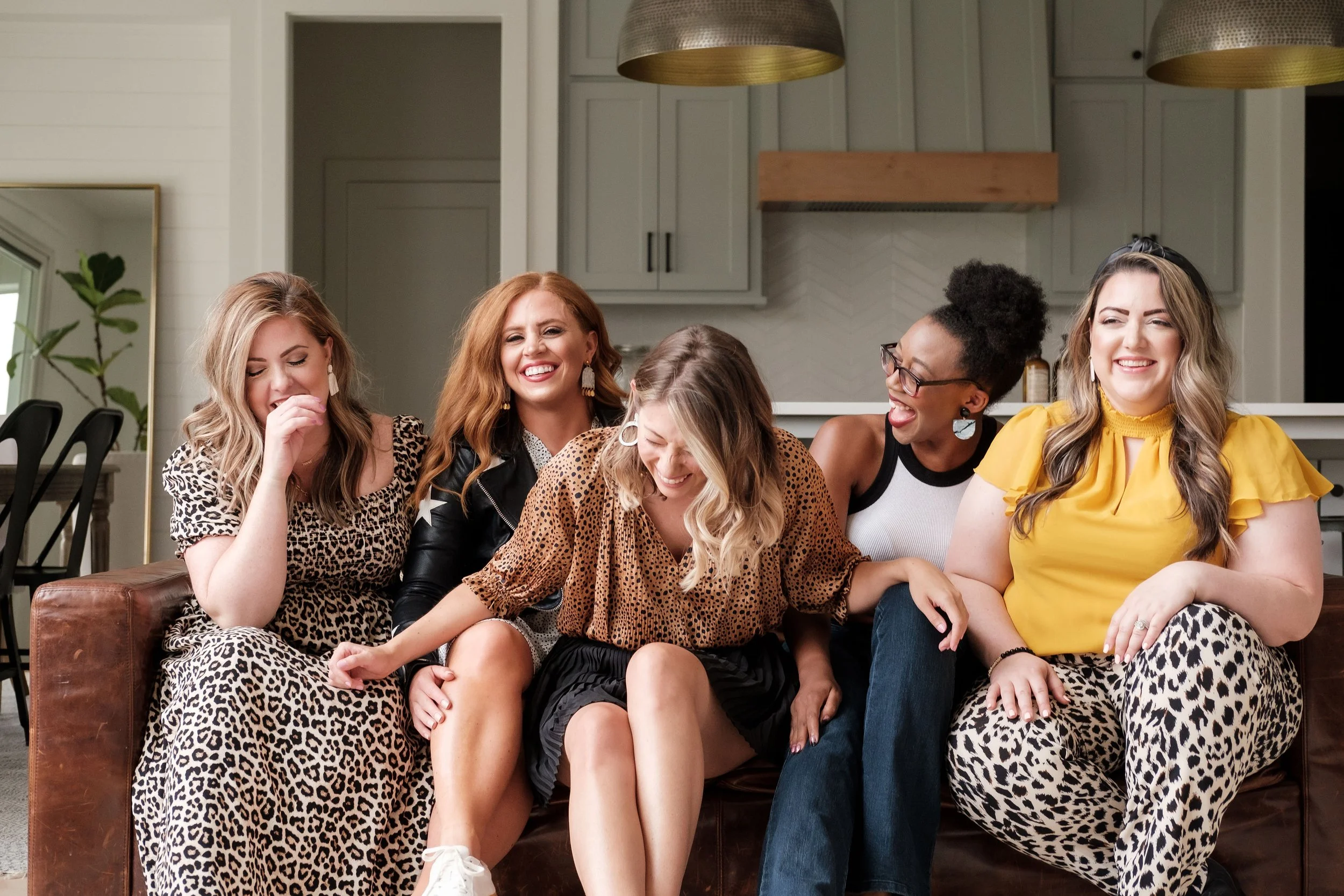 Five women sitting on a leather couch in a modern kitchen, laughing and enjoying each other's company.