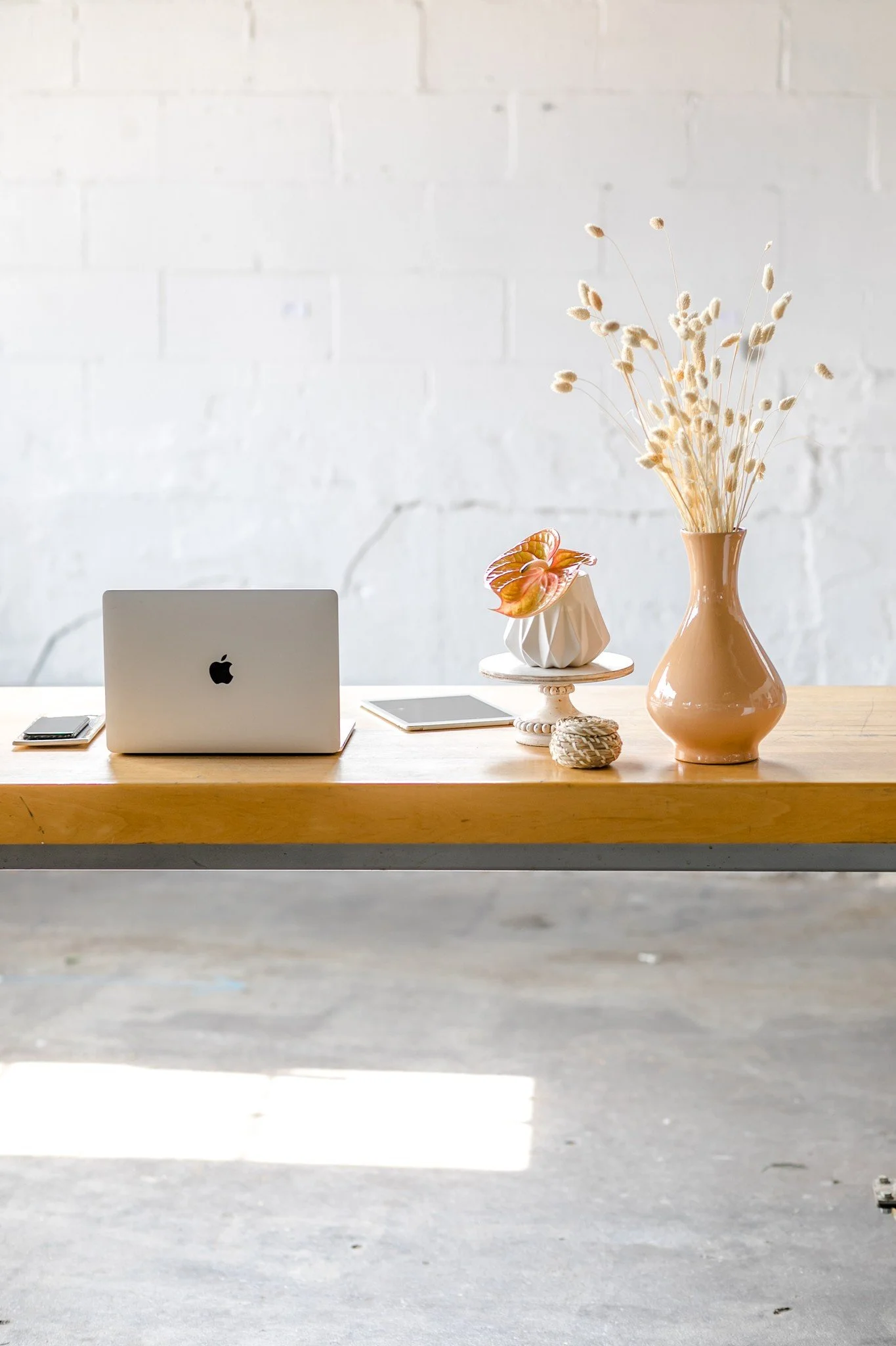 A wooden table with a silver Apple MacBook, a tablet, and a phone arranged on it. There is a decorative vase with dried flowers, a ceramic pot with an flower, a small decorative ball, and a small white pedestal with more dried flowers. The background is a white brick wall.
