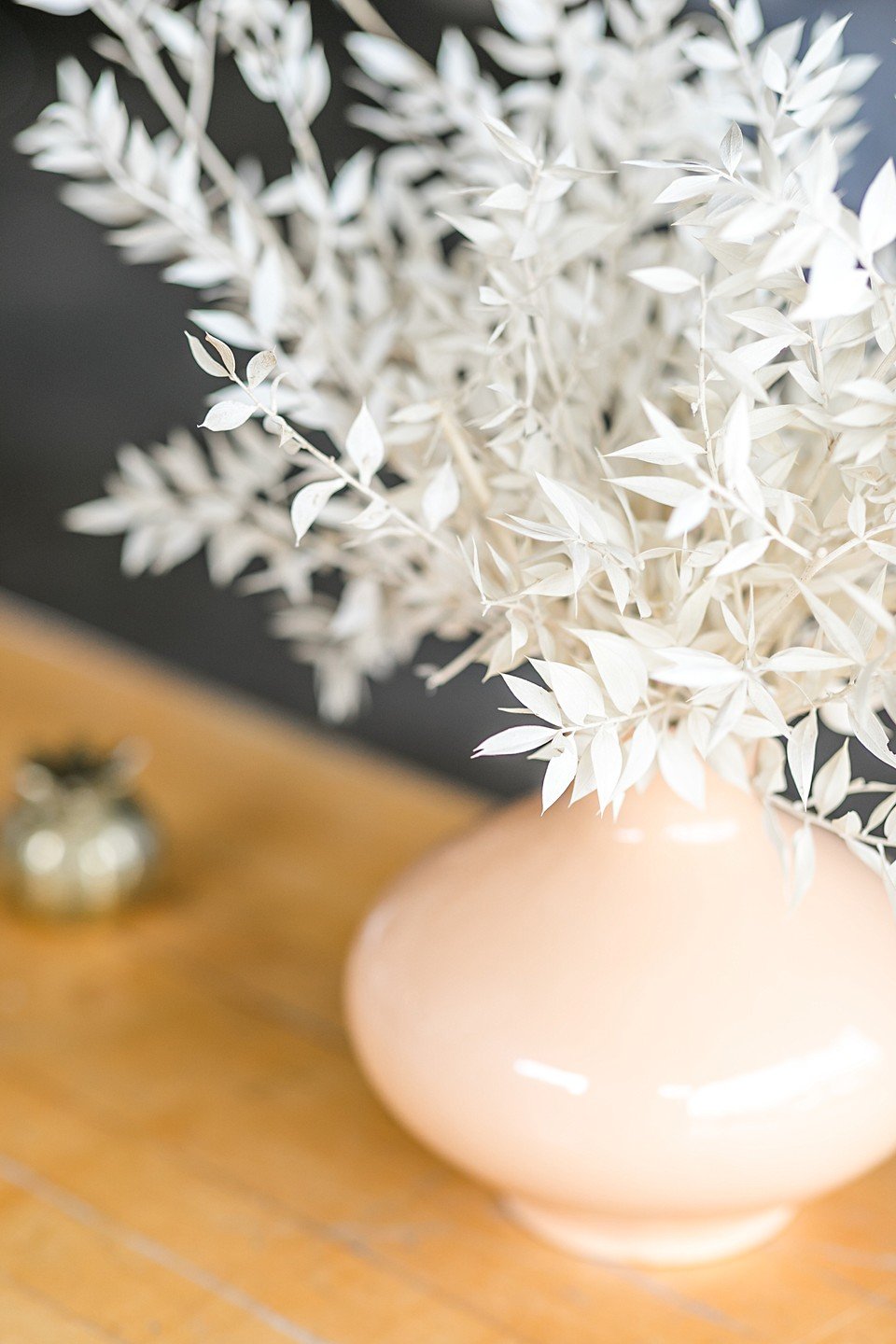 A beige ceramic vase with a glossy finish holding white-tinted dried foliage on a wooden surface.
