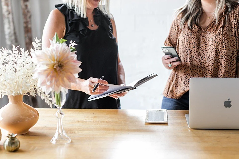 Two women standing at a wooden table, one holding a notebook and pen, the other looking at her phone, with a laptop, a smartphone, and a vase of flowers in front of them.