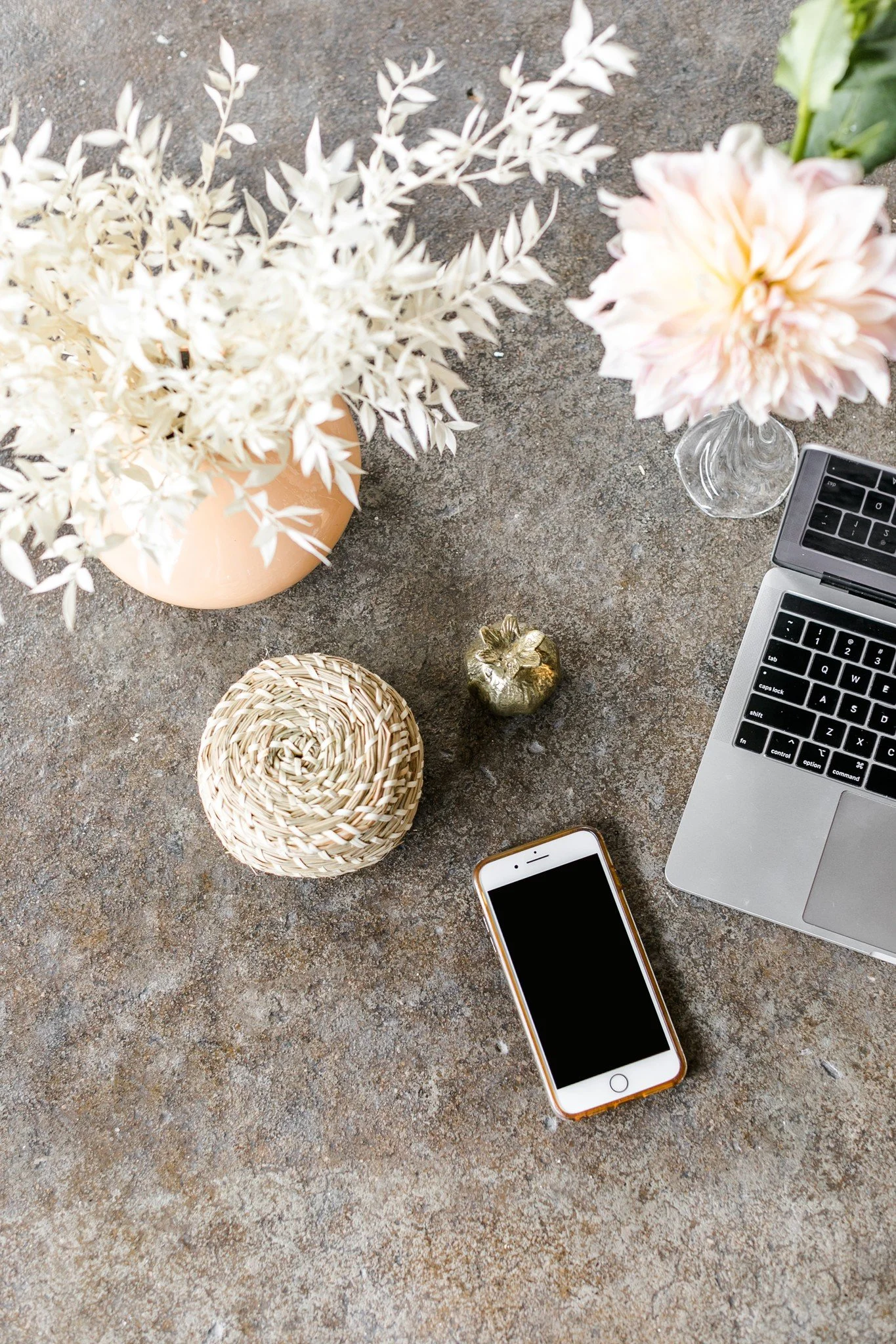 A flat lay of a workspace on a textured surface, featuring a laptop, a smartphone, a small gold pineapple figurine, a round woven container, and two vases with pink and white flowers and white foliage.