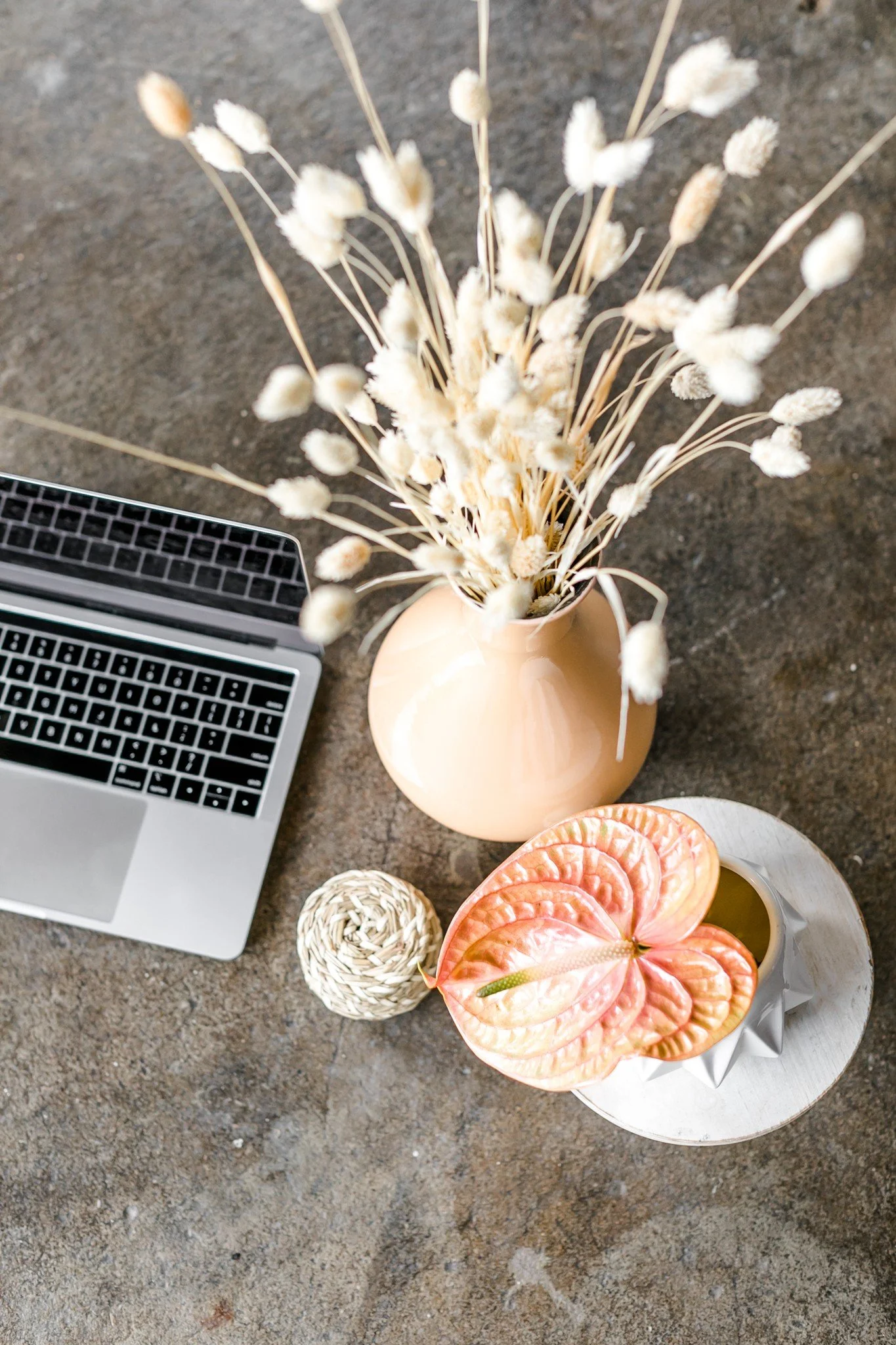 A flat lay of a workspace on a concrete surface includes a silver laptop keyboard, a beige vase with dried white fluffy flowers, a small spherical decorative object, and a white geometric vase with pink and green leaves, all arranged with a white round tray.