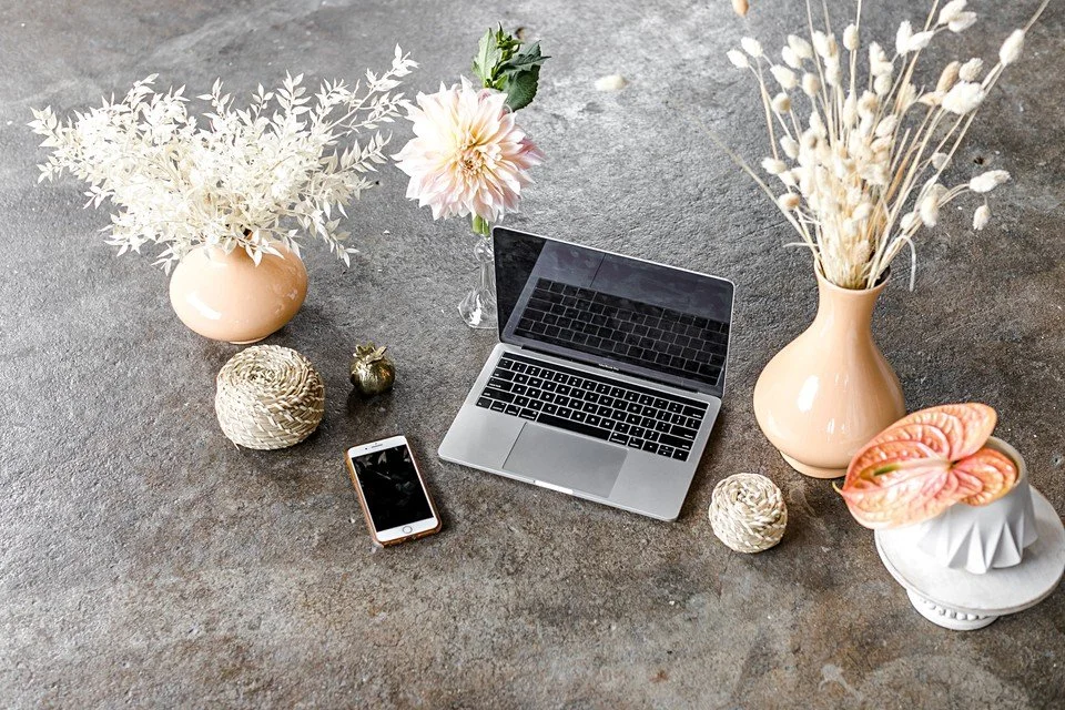 A laptop, a smartphone, and decorative vases with flowers and plants arranged on a gray surface.