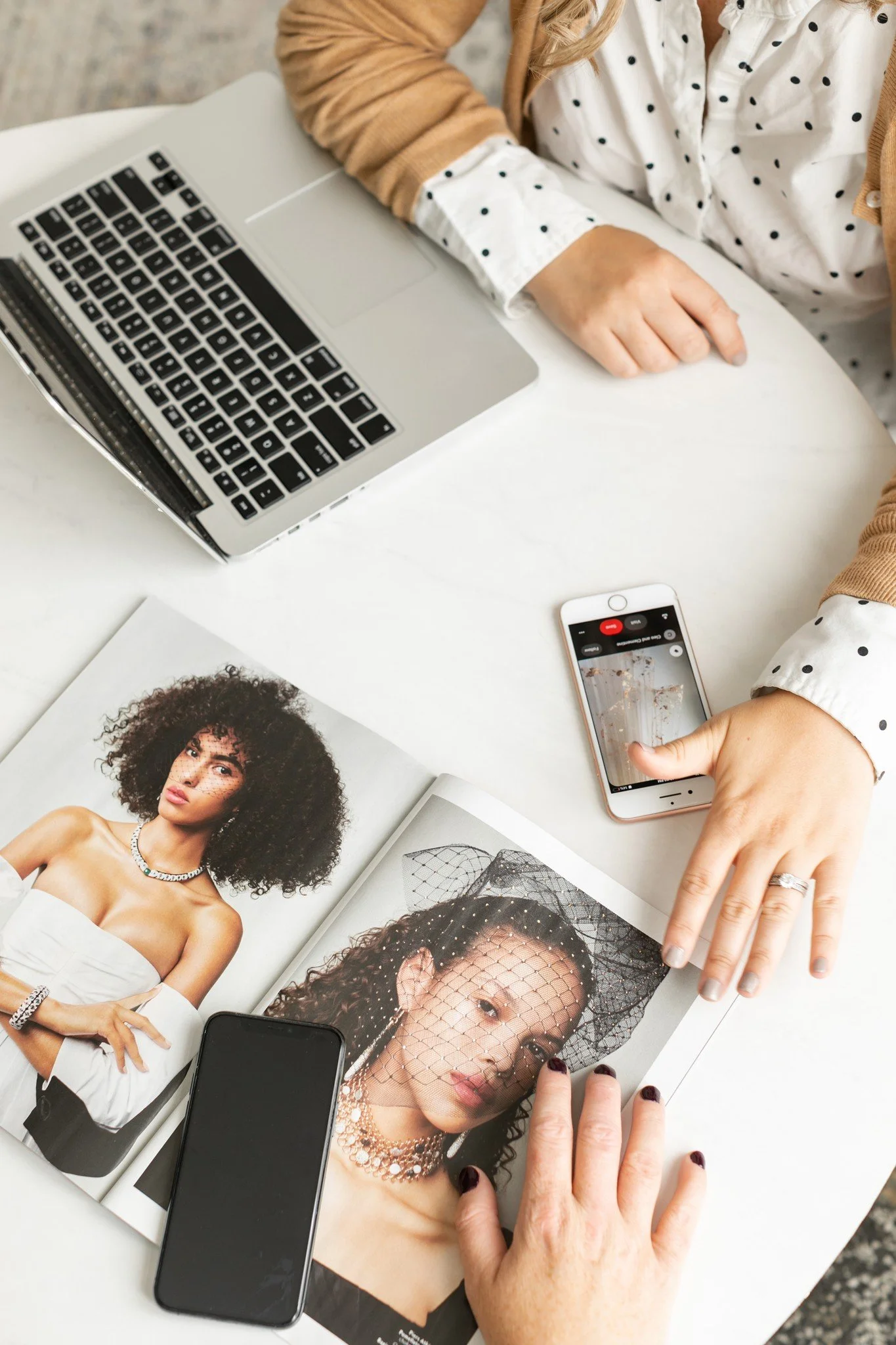 A person working on a laptop and reviewing fashion magazines with model photos, while using two smartphones on a white table.