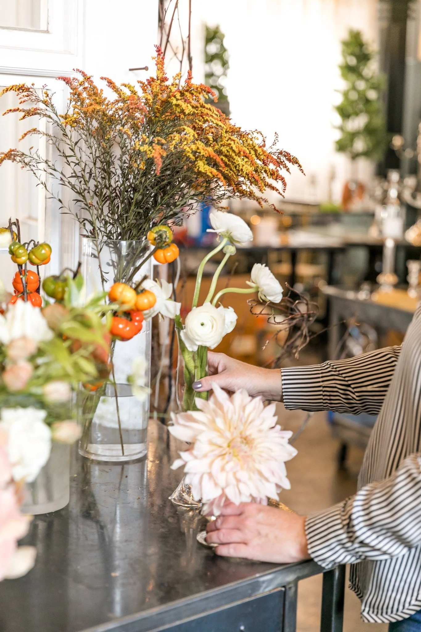 Person arranging white and pale pink flowers in a glass vase on a black table, surrounded by other colorful flower arrangements.