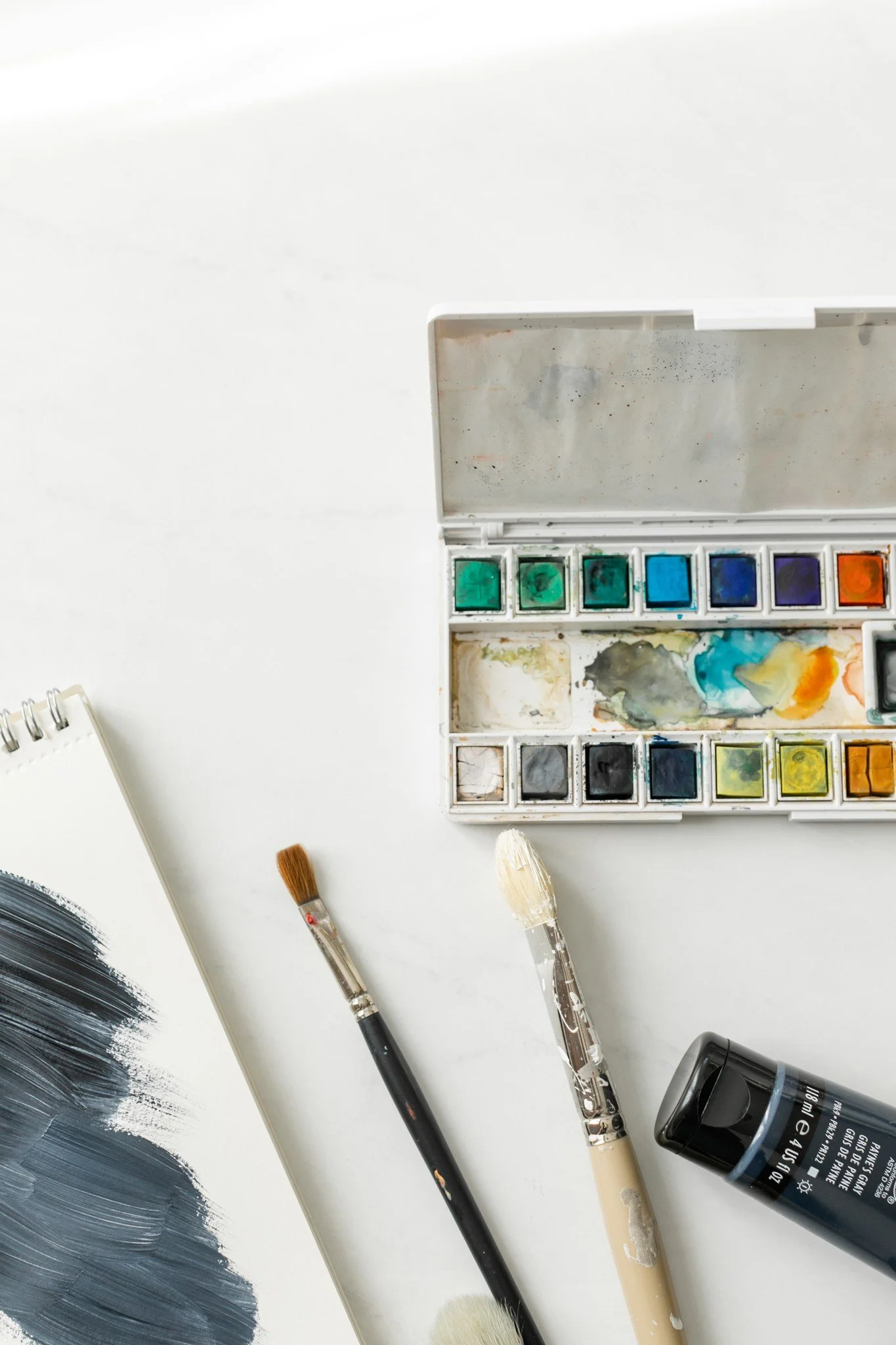 Watercolor paints, brushes, and a palette on a white surface, with a partially painted paper and a black bottle of paint or ink.