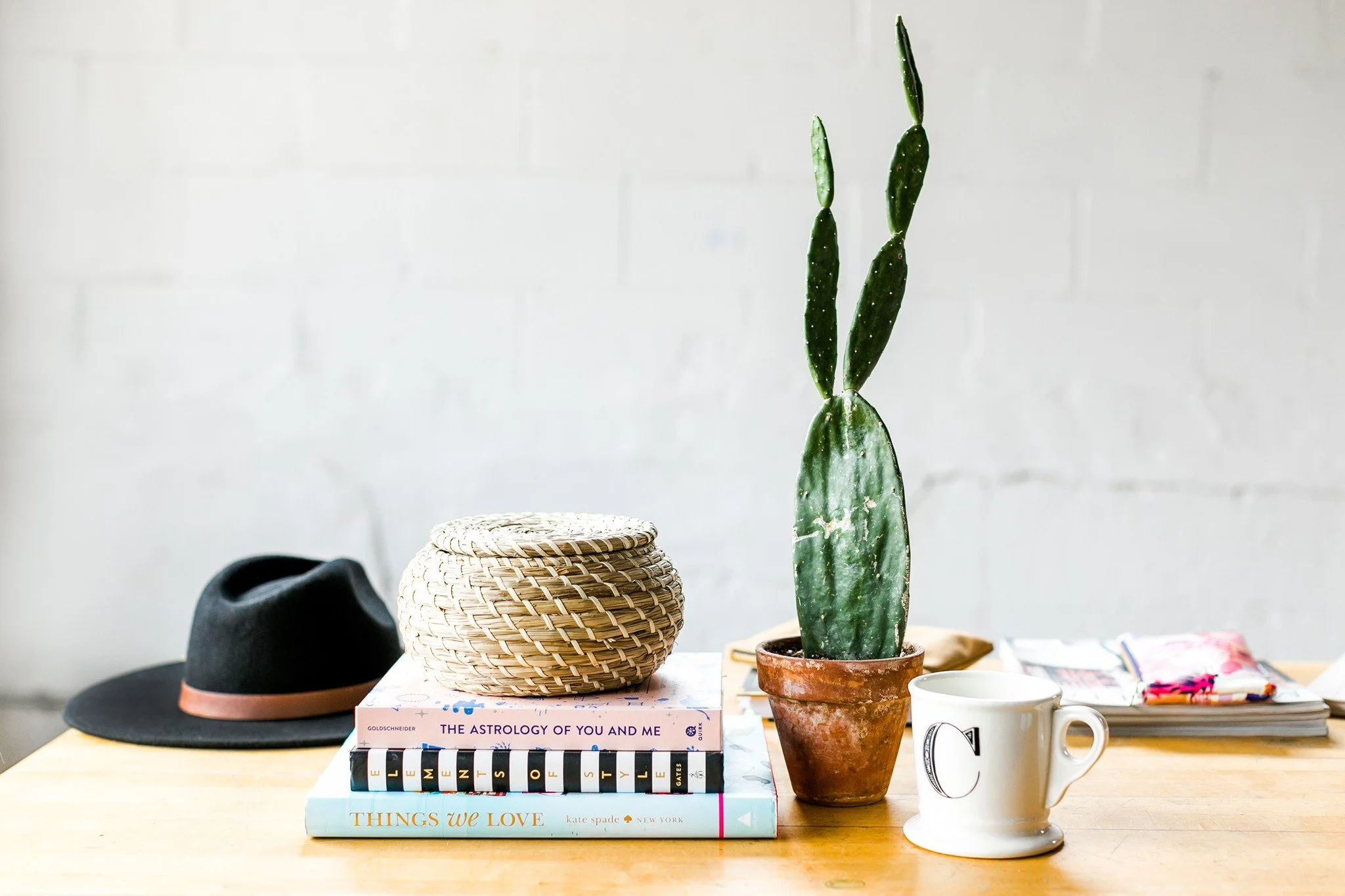A wooden table with a black hat, a woven basket, three books titled "The Astrology of You and Me," "Element," and "Things We Love," a potted cactus plant, a white mug with a black letter "C," and various items partially visible on the right, against a white brick wall.
