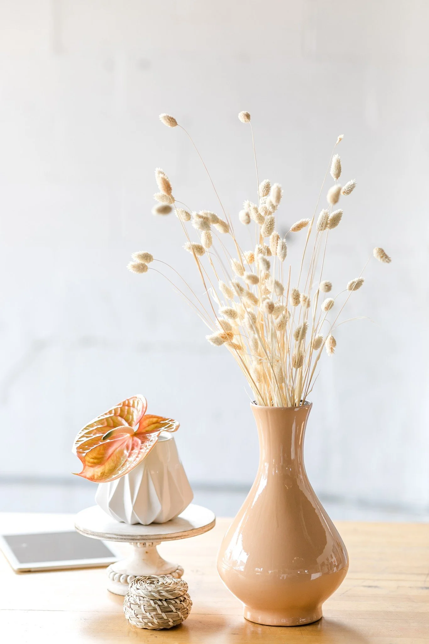 A beige ceramic vase with dried tall grass on a light wooden table. There is a white decorative object on a small pedestal, a small round woven ball, and a white tray with a tablet on the table.