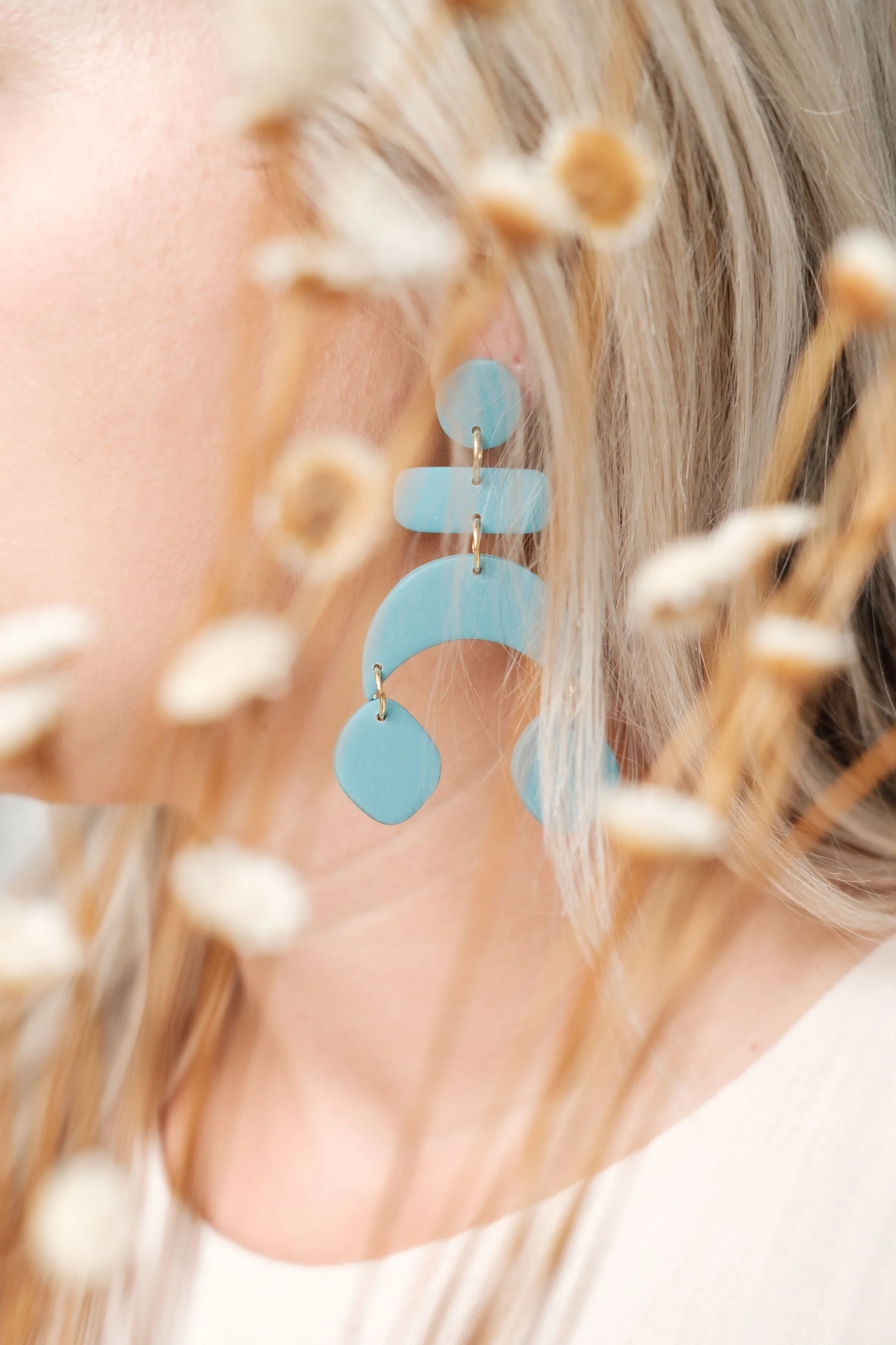 Close-up of a woman wearing a large, light blue, geometric earring, surrounded by blurred dried flowers.