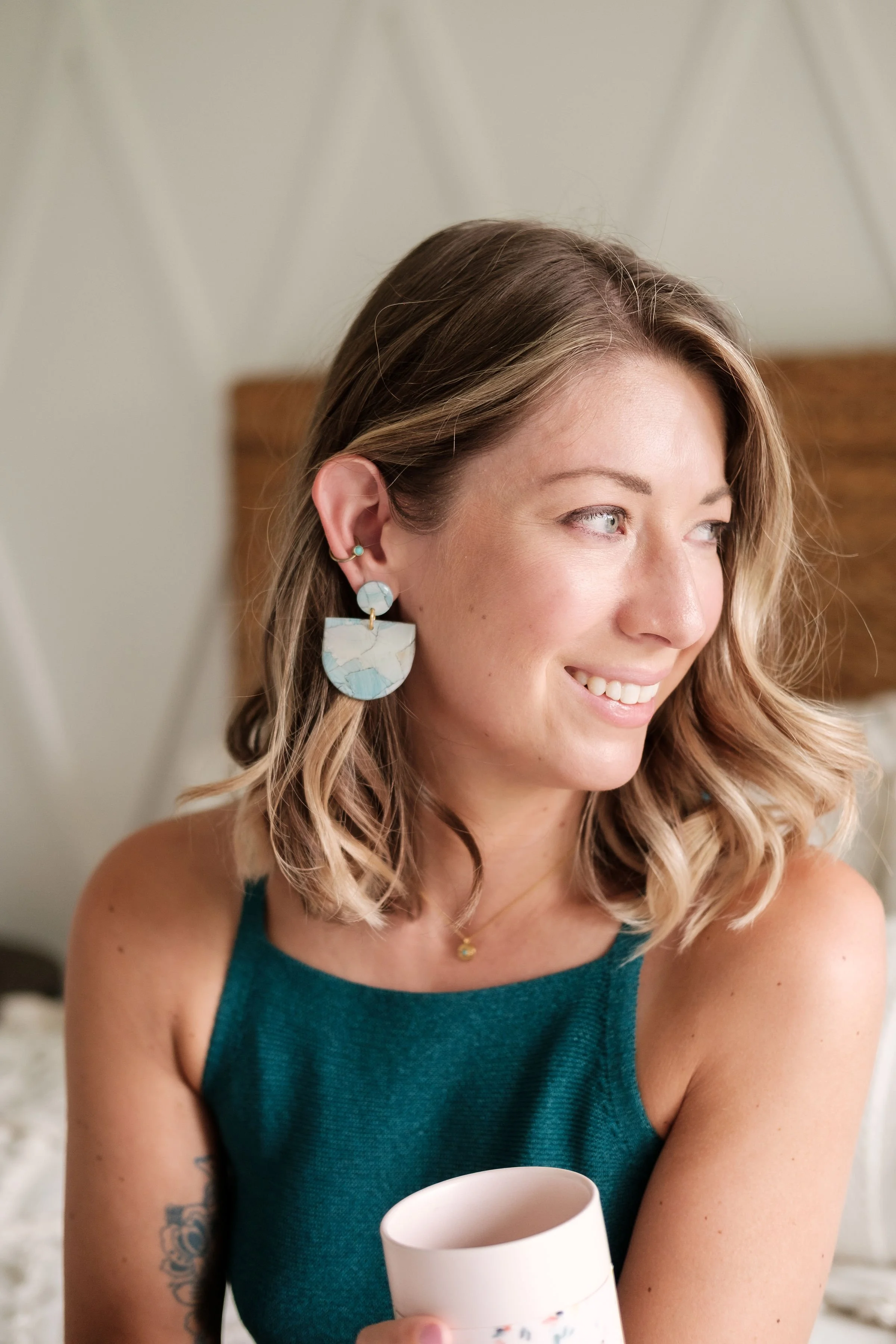 A woman with wavy blonde hair smiling, holding a white mug. She is wearing a teal sleeveless top, and large earrings with marble-like patterns, along with a delicate necklace. The background shows a wooden headboard and light-colored walls.