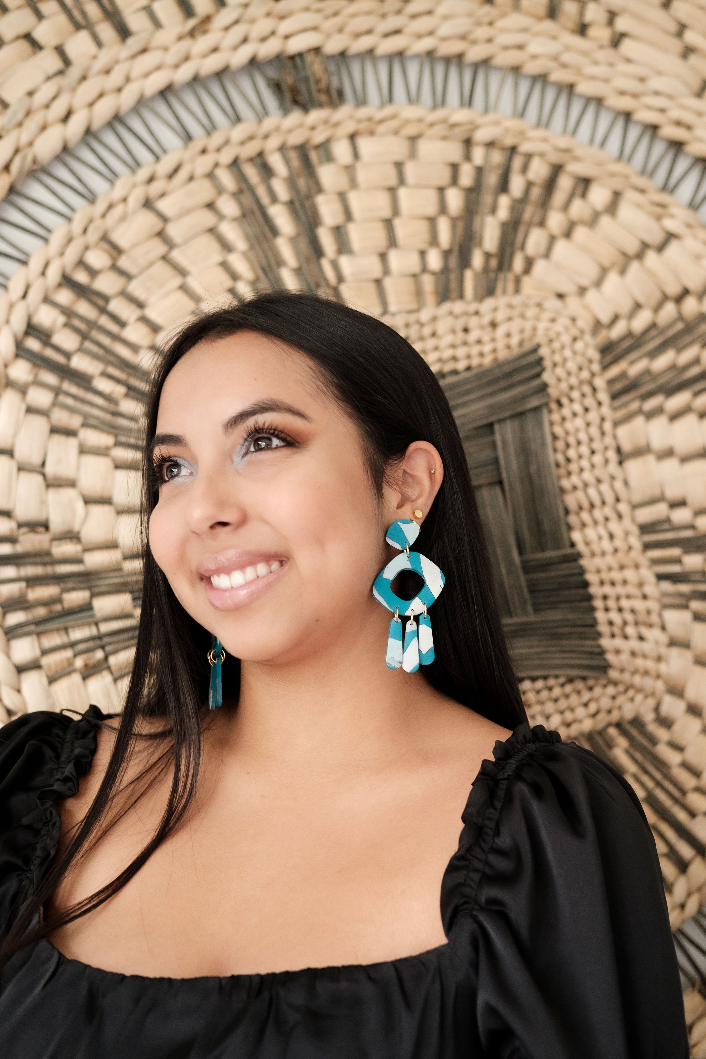 A woman with long dark hair smiling, wearing black top and large colorful earring, standing in front of a circular woven wall decor.