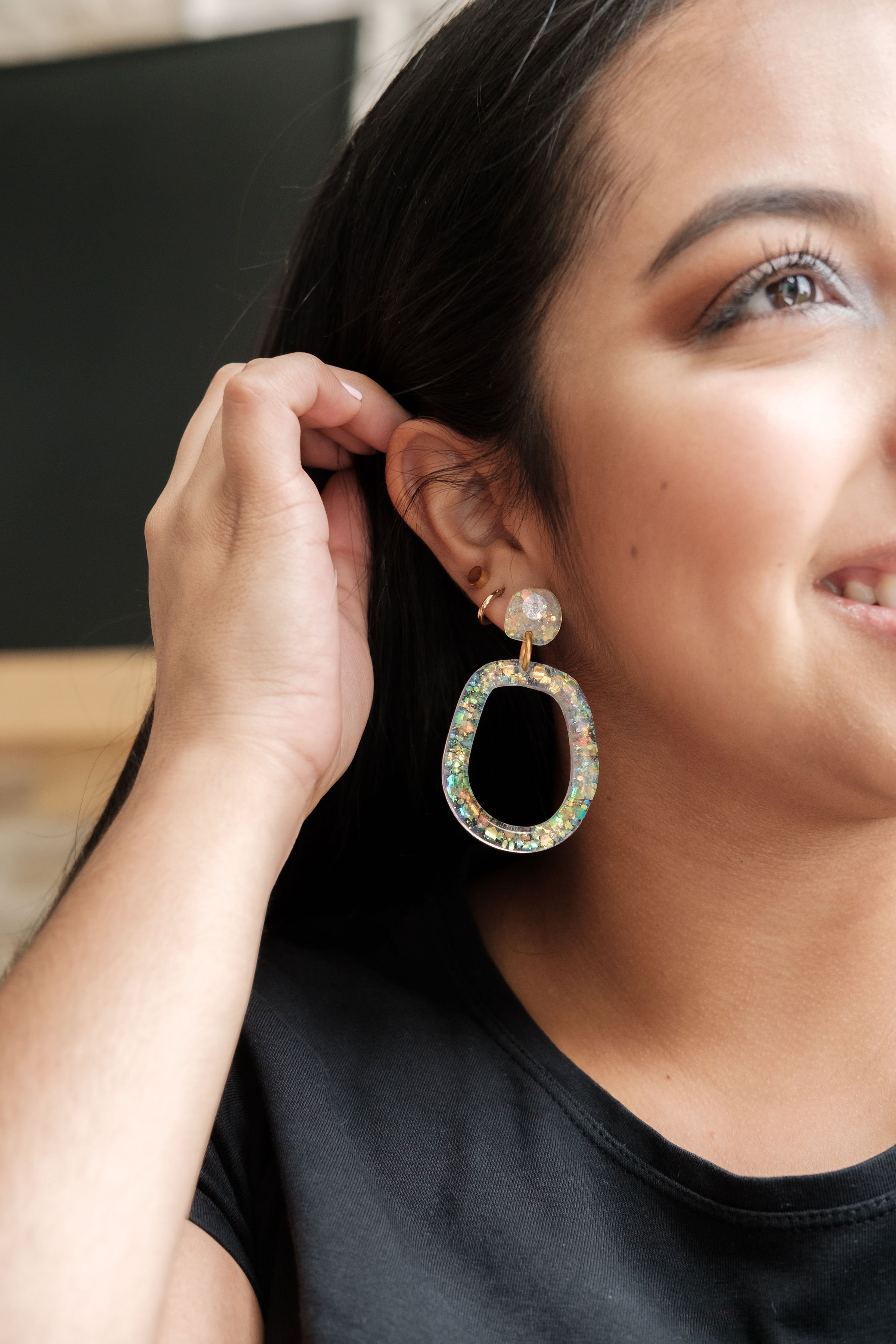 A woman wearing a large iridescent earring that has a teardrop shape with a circular top, and a small gold hoop in her ear.