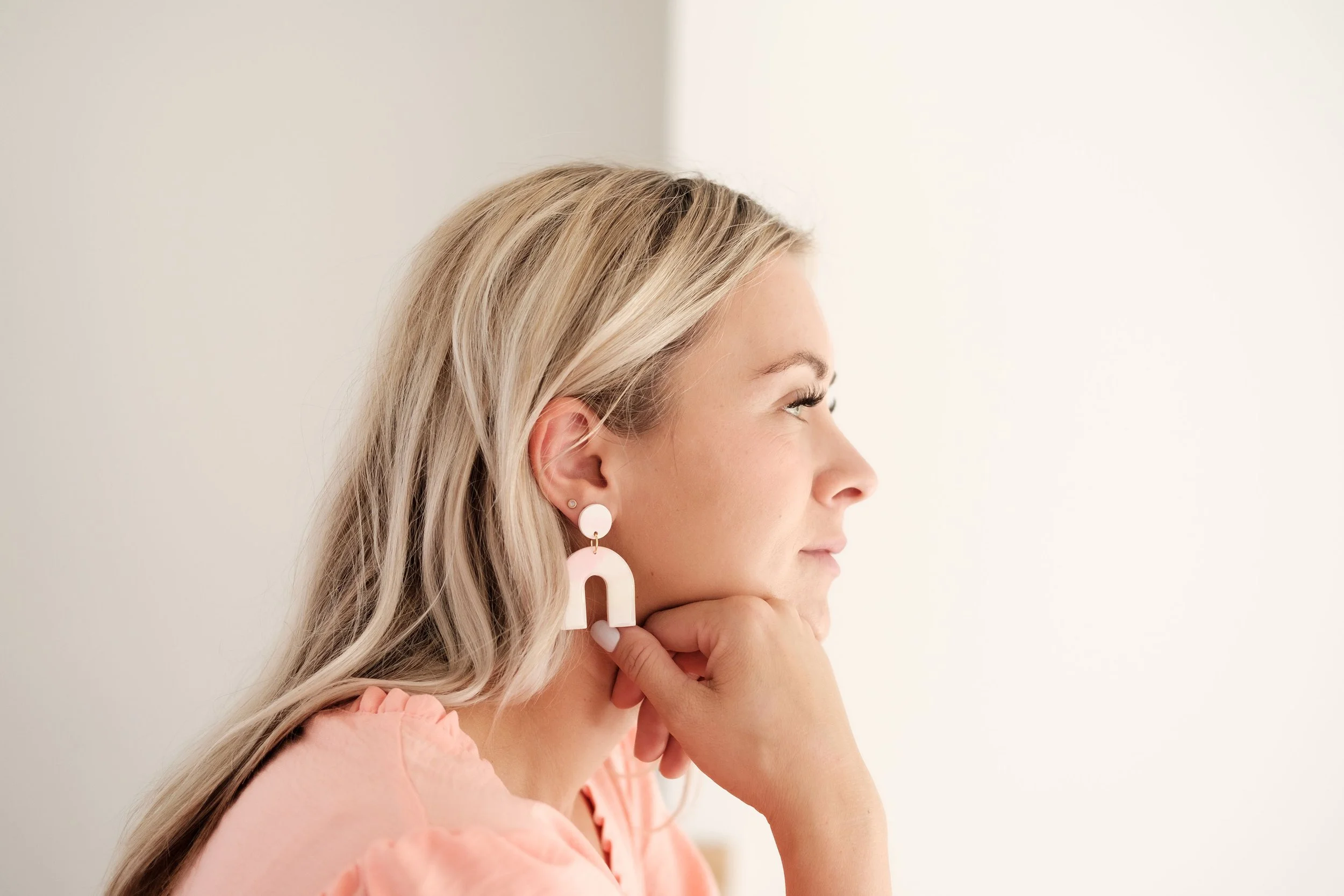 Side profile of a smiling blonde woman wearing large pink hoop earrings and a pink top, resting her chin on her hand against a plain background.