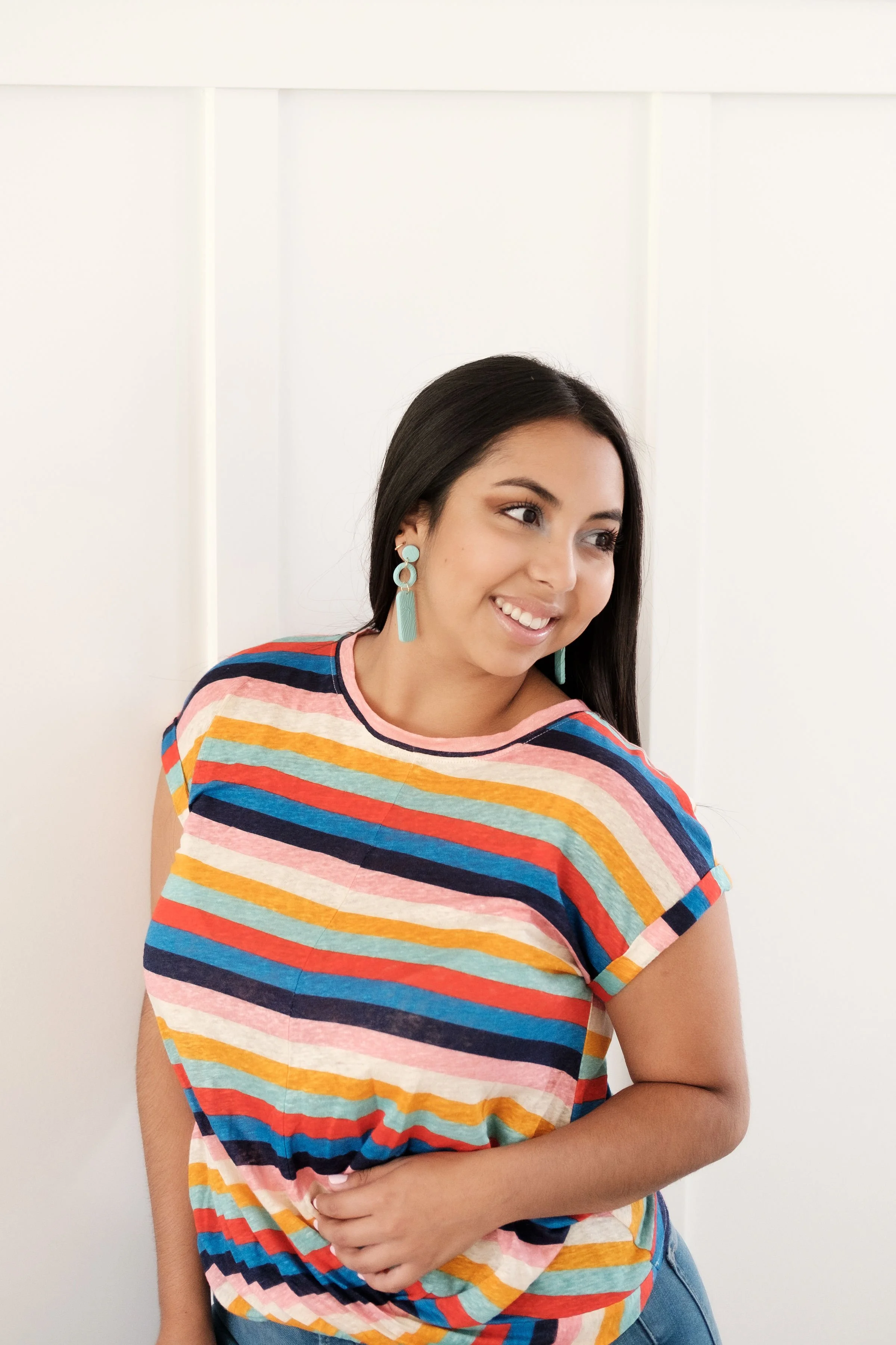 A young woman with long dark hair, wearing teal earrings and a multicolored striped T-shirt, smiling and looking to her right, standing against a white paneled wall.