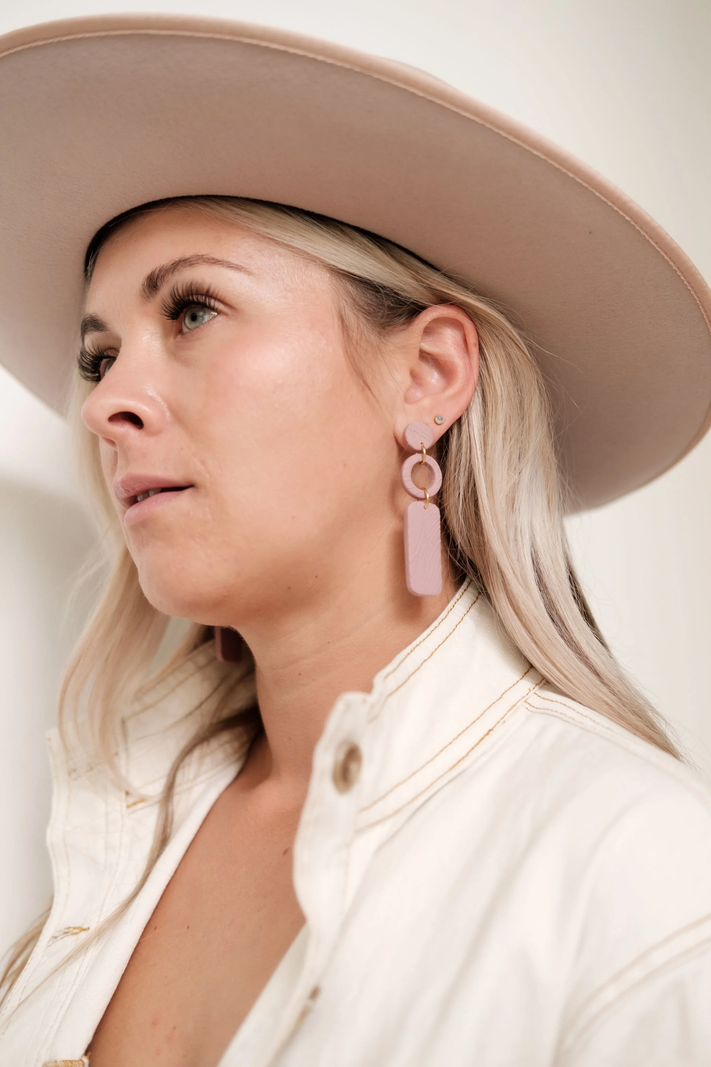 A woman wearing a beige wide-brimmed hat, pink earrings, and a cream-colored shirt with a button near the collar, looking to her left.