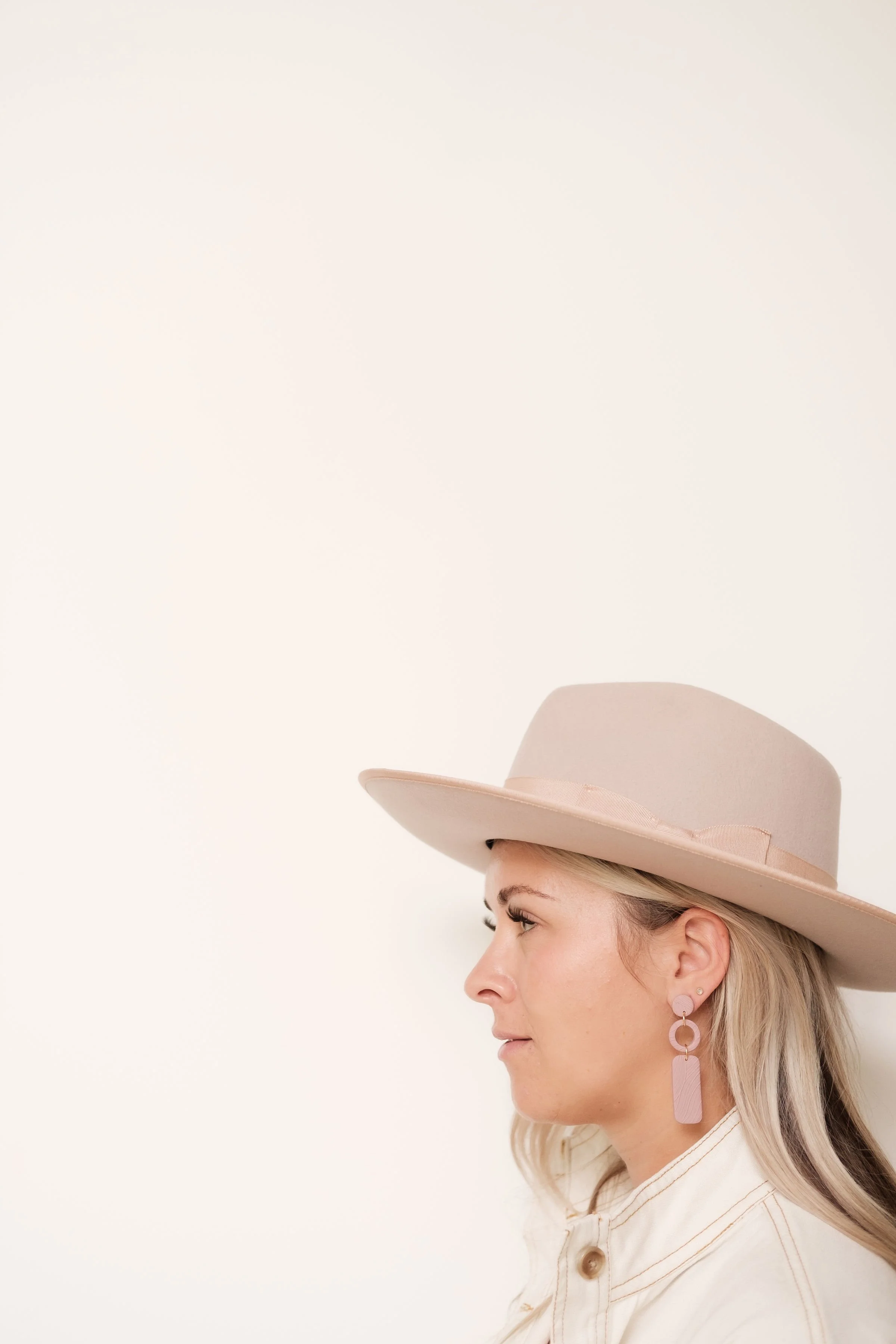 Profile of a woman with blonde hair wearing a wide-brimmed pink hat and pink earrings, against a plain white background.