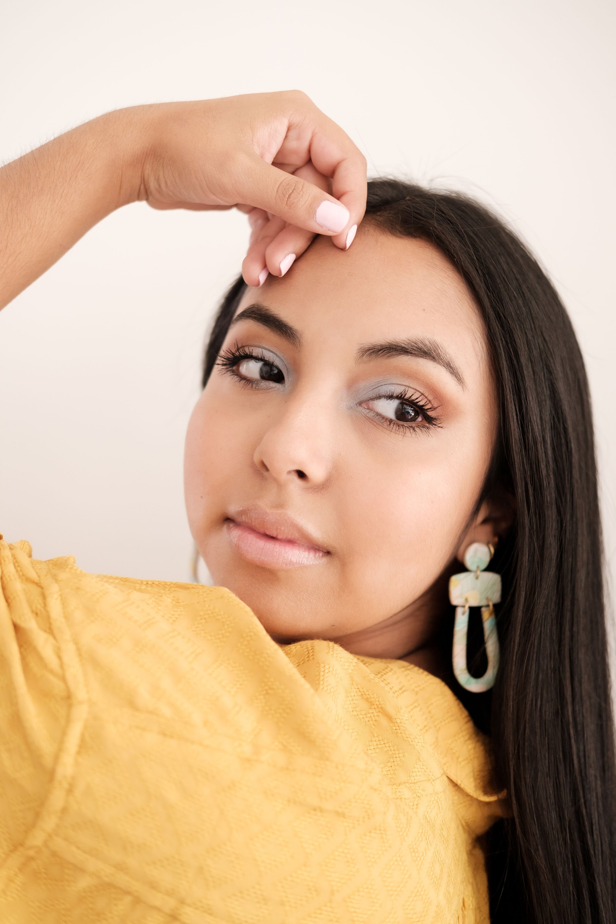 Close-up of a young woman with long dark hair wearing a yellow textured top and large earrings, with her hand resting on her forehead and looking to the side.