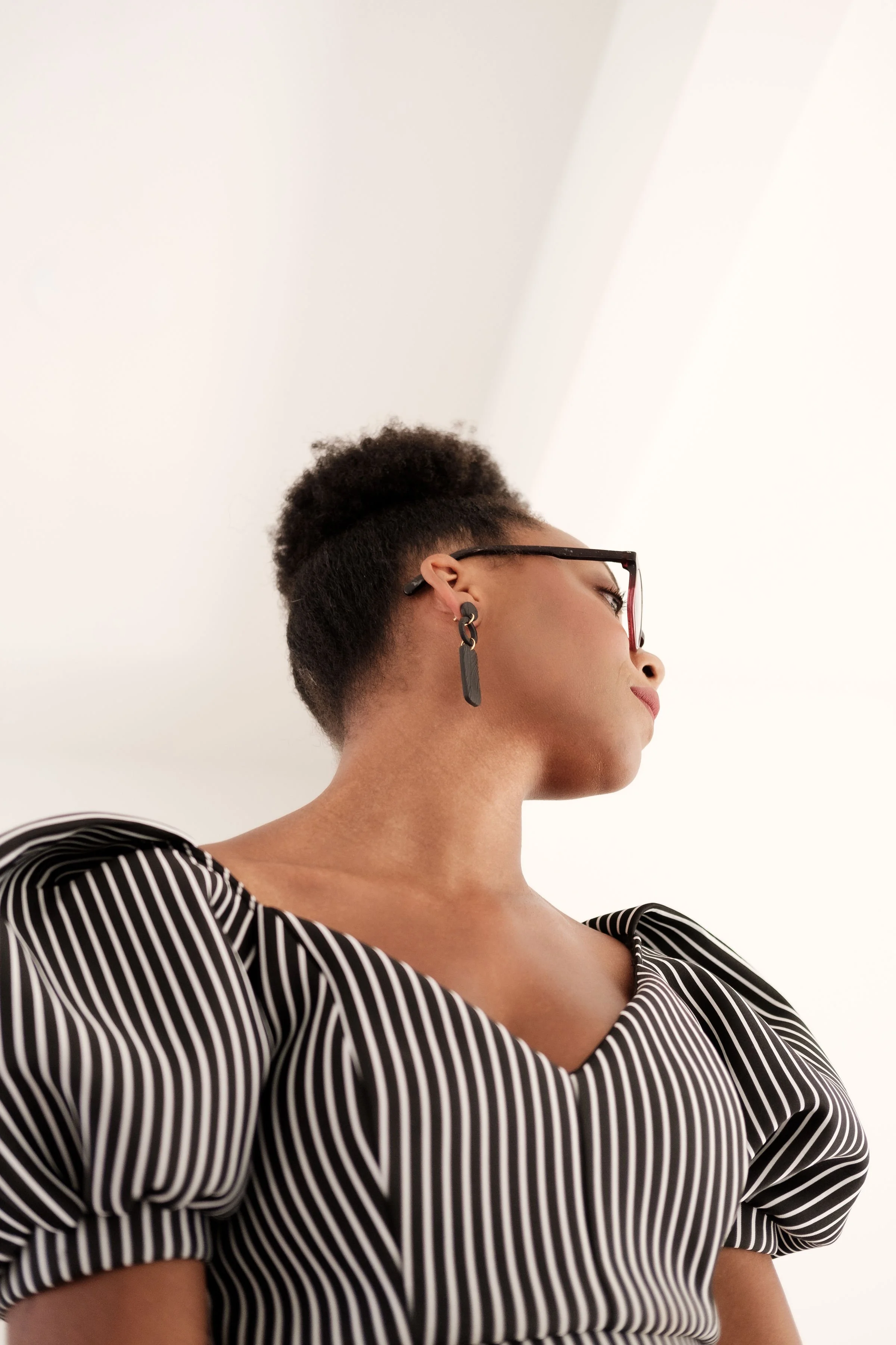 Side profile of a woman with short curly hair wearing glasses, black and white striped top, and black earrings, standing against a white ceiling background.